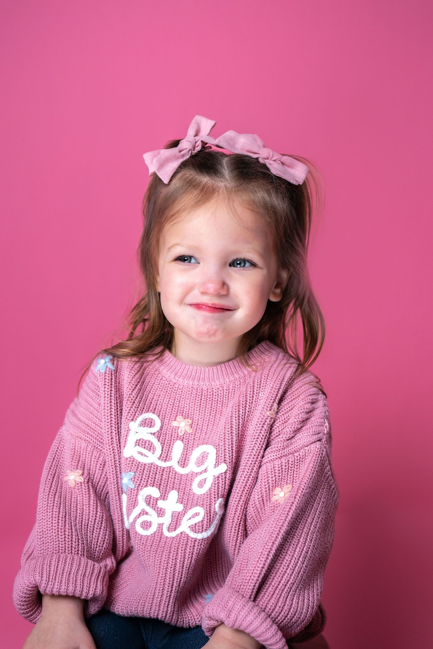 A child wearing a pink knitted sweater with "Big Sis" text and matching hair bows poses against a pink background.
