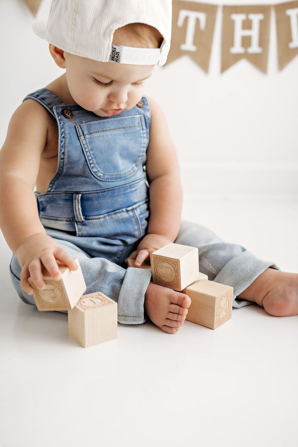 A small child in denim overalls and white hat sits on the floor playing with wooden toy blocks.