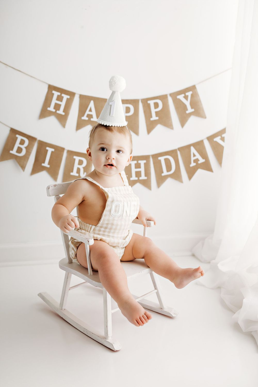 A baby in white romper sits on a rocking chair with Happy Birthday burlap banner hanging behind against white backdrop.