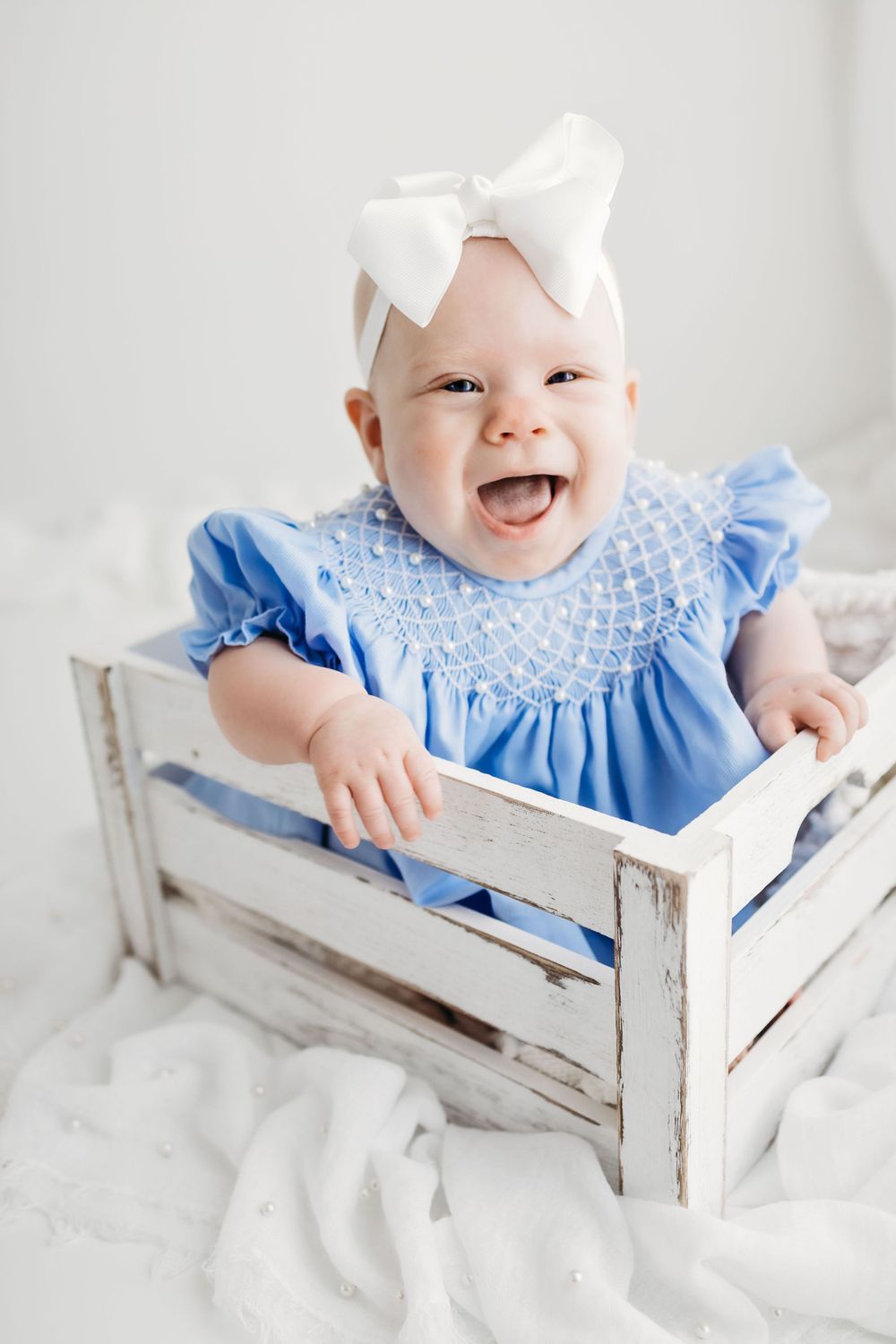 A joyful baby wearing a blue smocked dress and white bow sits in a distressed white wooden crate.