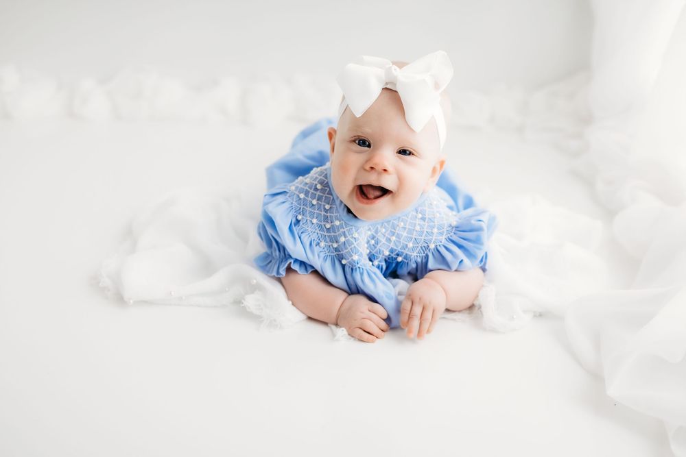 Small baby wearing blue ruffled dress and white bow lying on white bedding with a joyful smile.