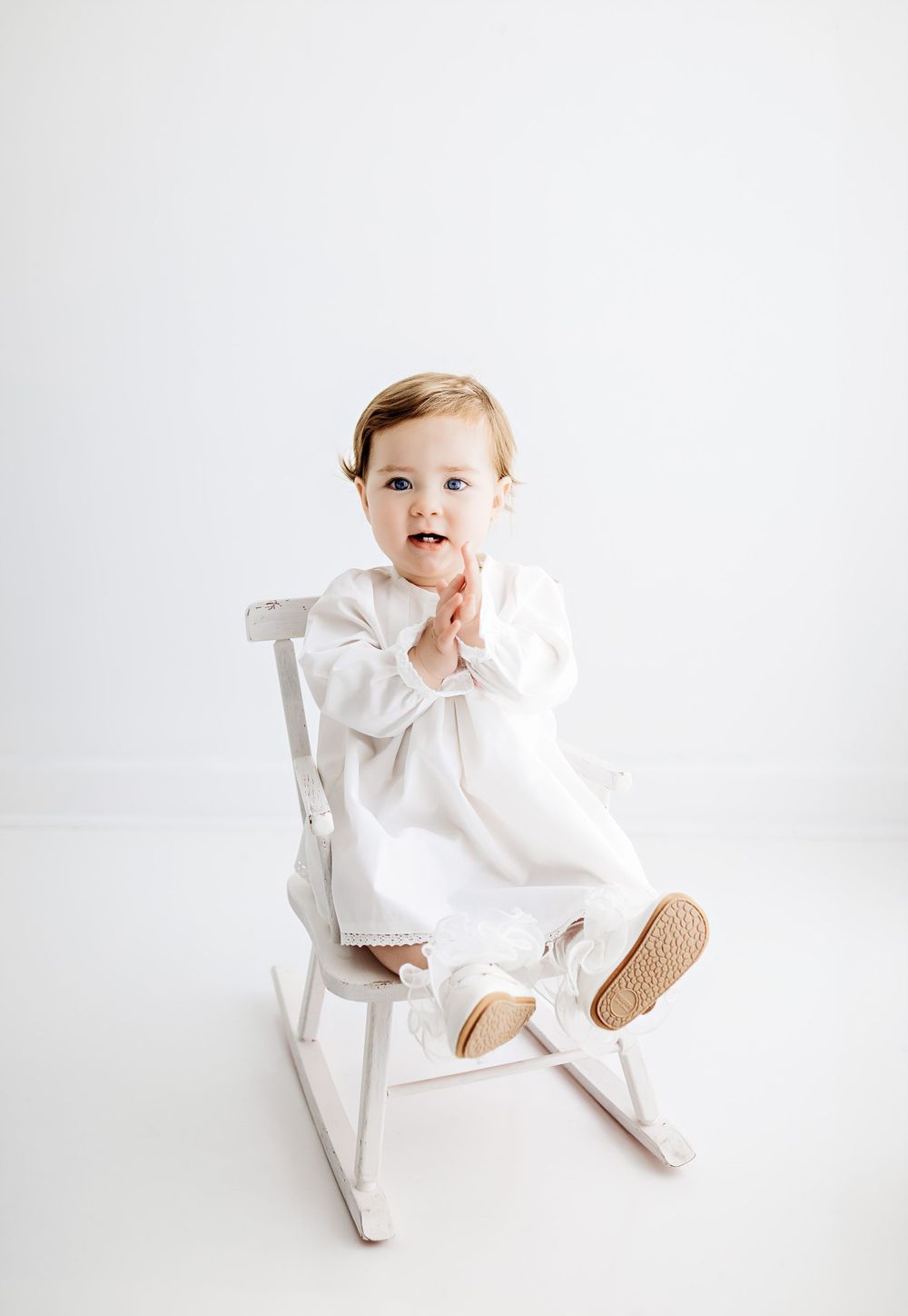 Small child in white dress and shoes sitting on a white rocking chair against light background.