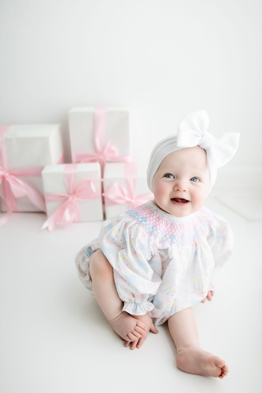 Baby in white smocked outfit and bow sits beside white gift boxes with pink ribbons against white background.