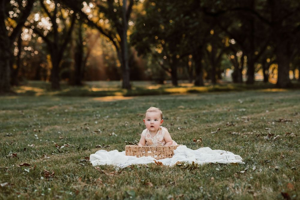 A toddler in a knit sweater sits on a white blanket in a scenic autumn park setting.