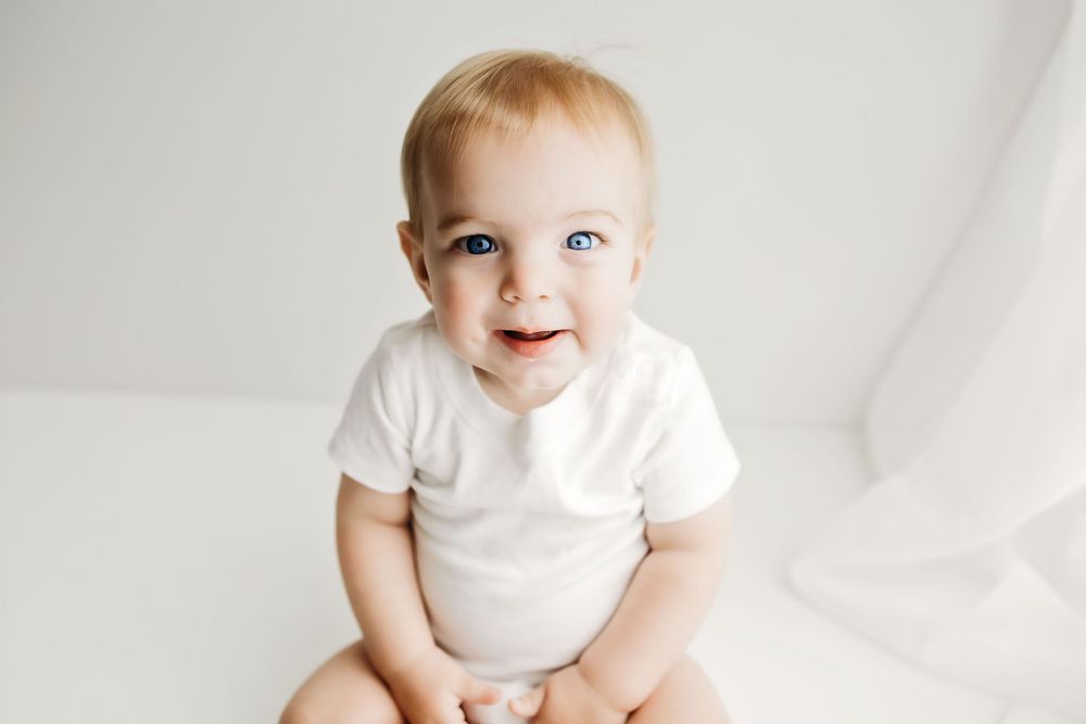 Smiling baby wearing white bodysuit sits against light background while looking up with bright blue eyes.