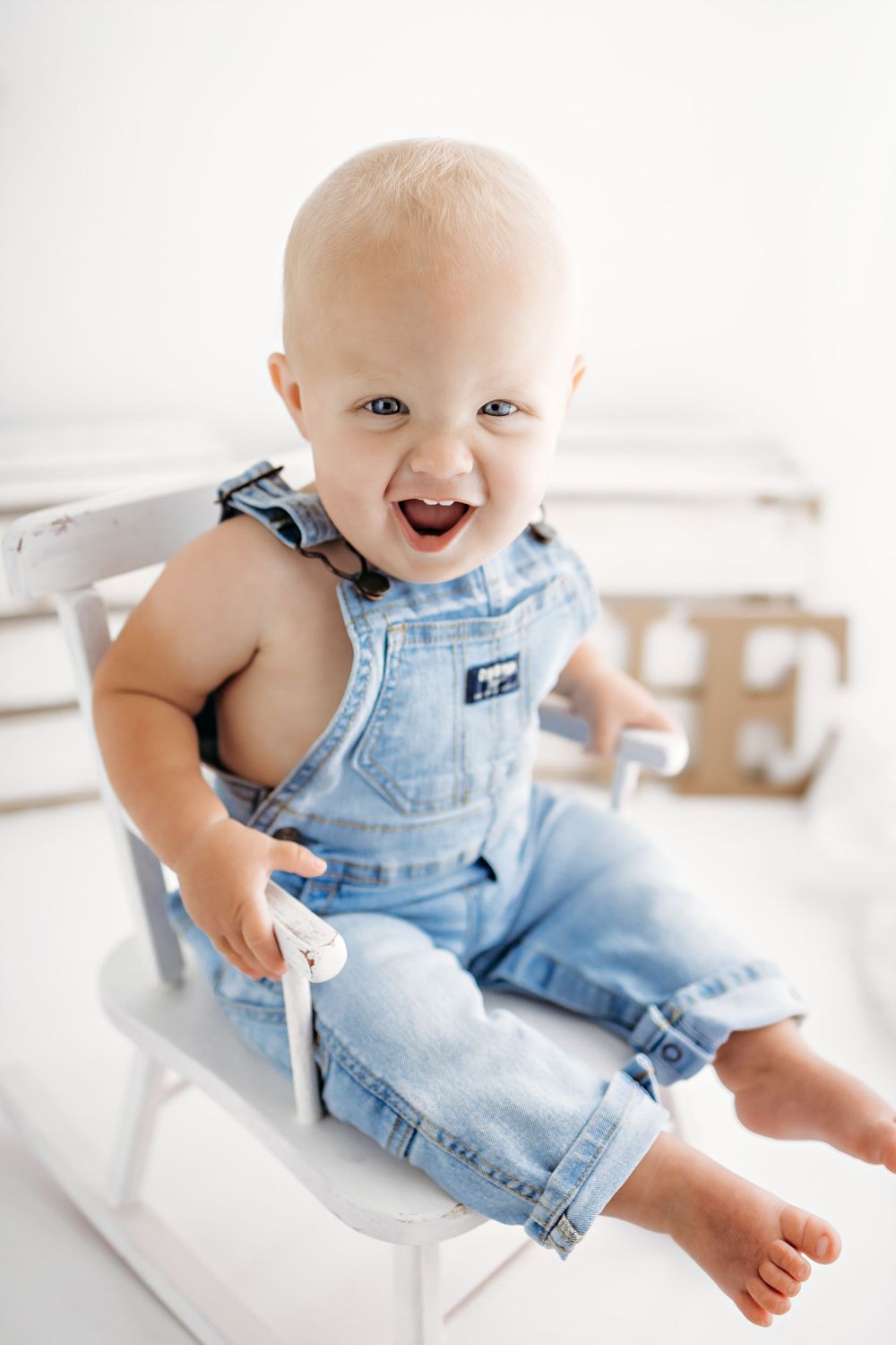 Happy baby wearing light blue denim overalls sits in white chair with bare feet and bright expression.
