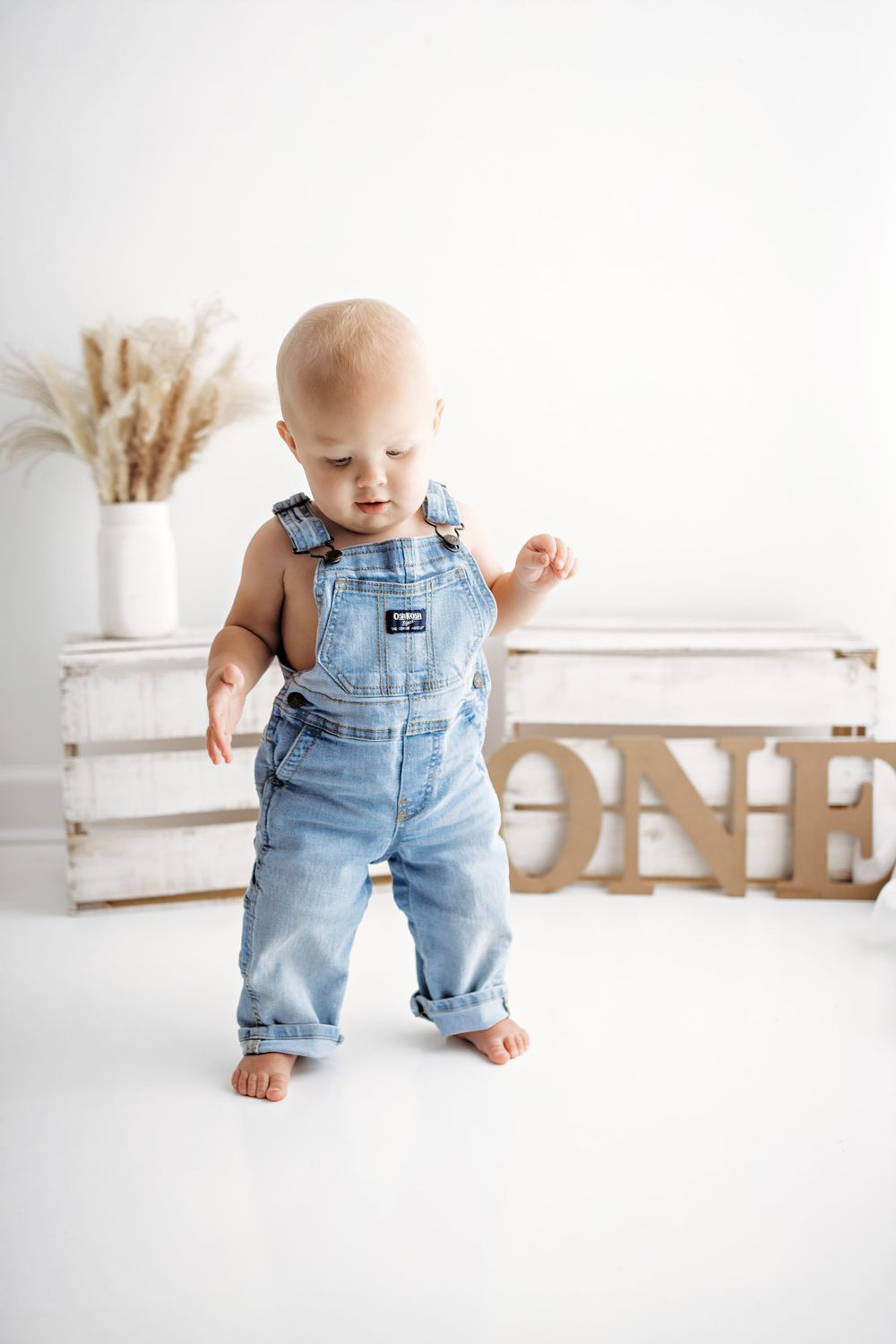 Baby wearing denim overalls stands barefoot near wooden crate and dried pampas grass decor against white backdrop.