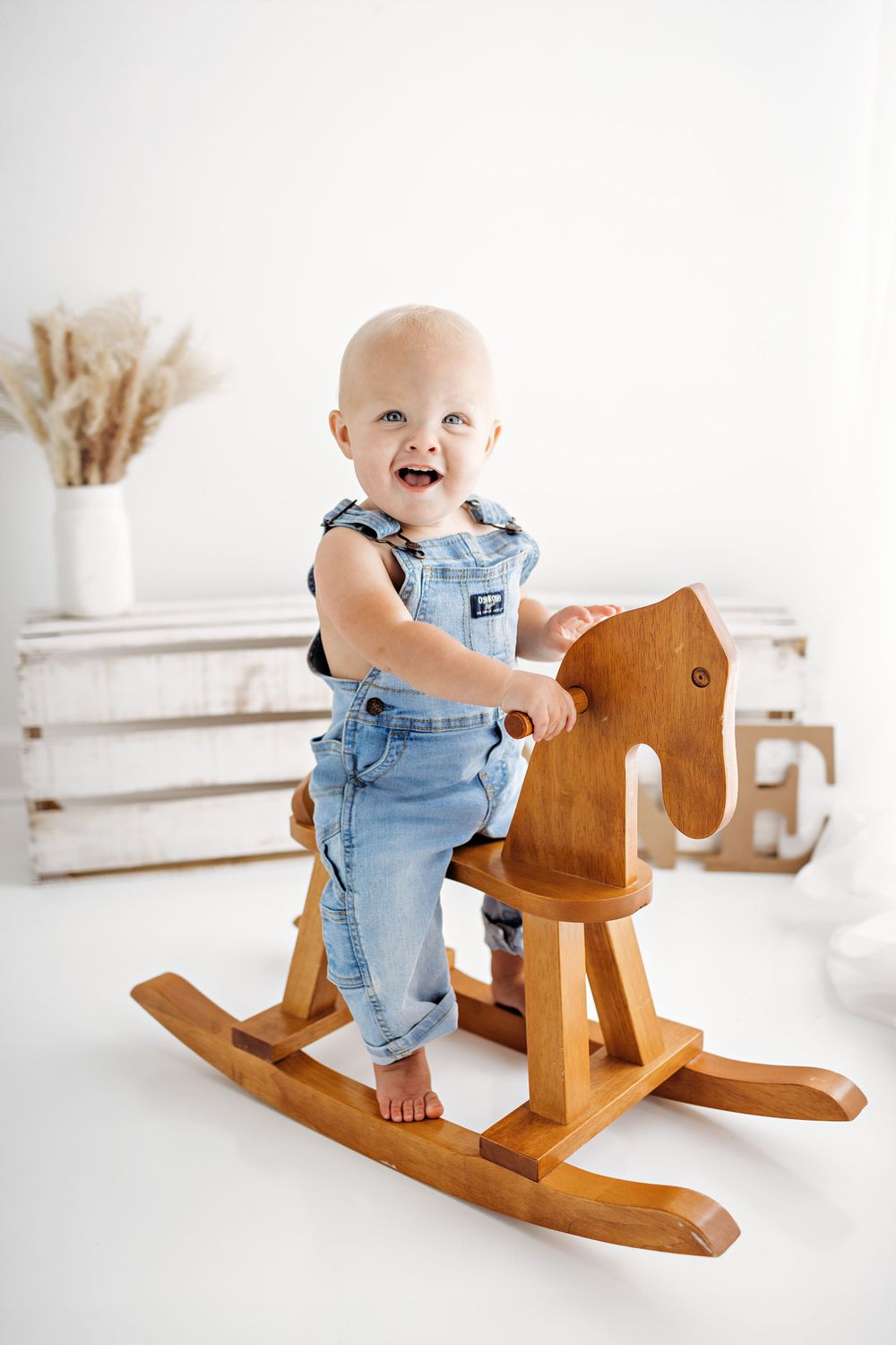 A small child in denim overalls stands and smiles while playing on a wooden rocking horse toy against a white backdrop.