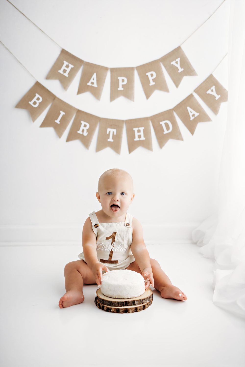 First birthday celebration setup with rustic burlap banner and white smash cake on wooden slice against white backdrop.