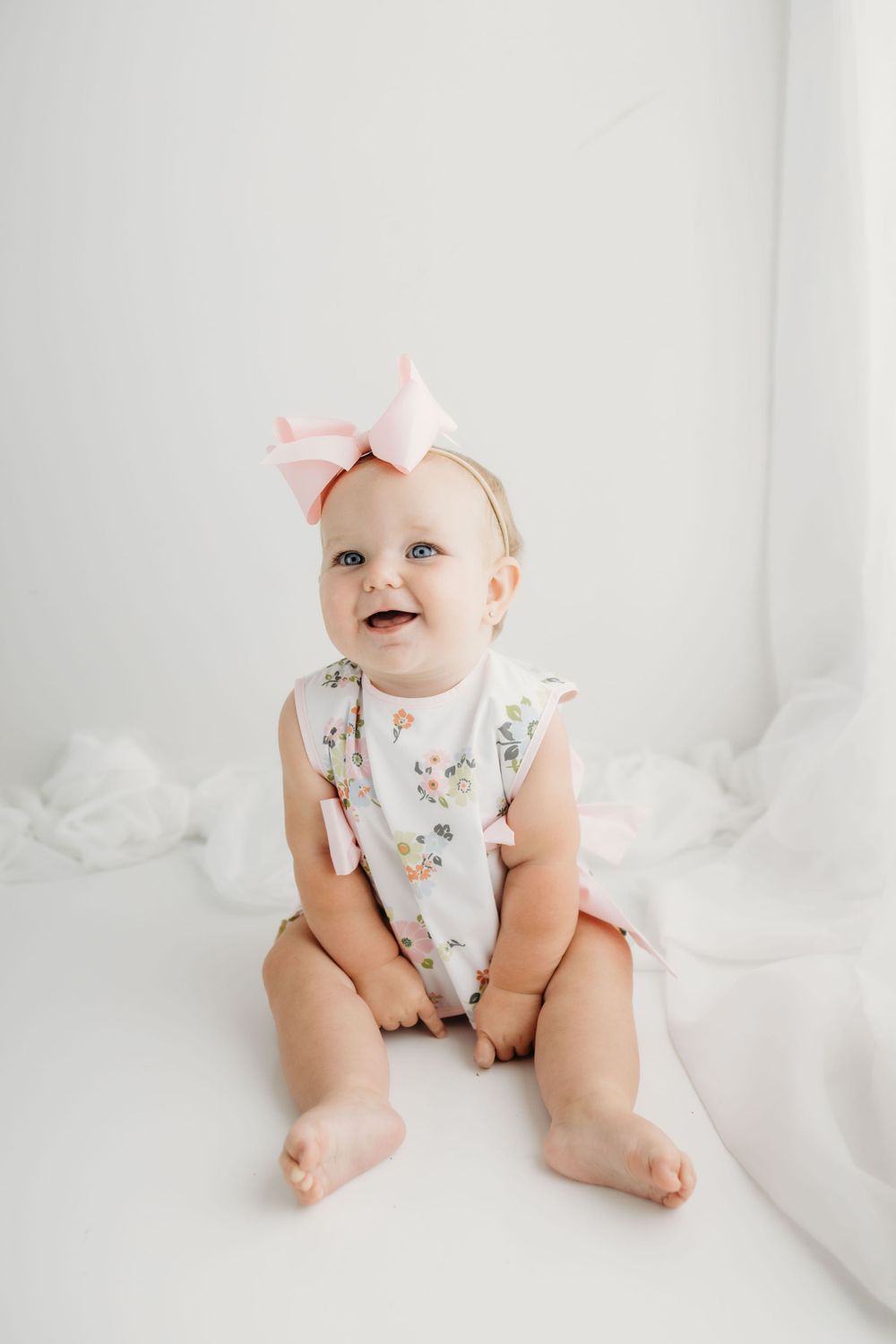 A baby wearing a floral romper and pink bow headband sits on white bedding while smiling joyfully.