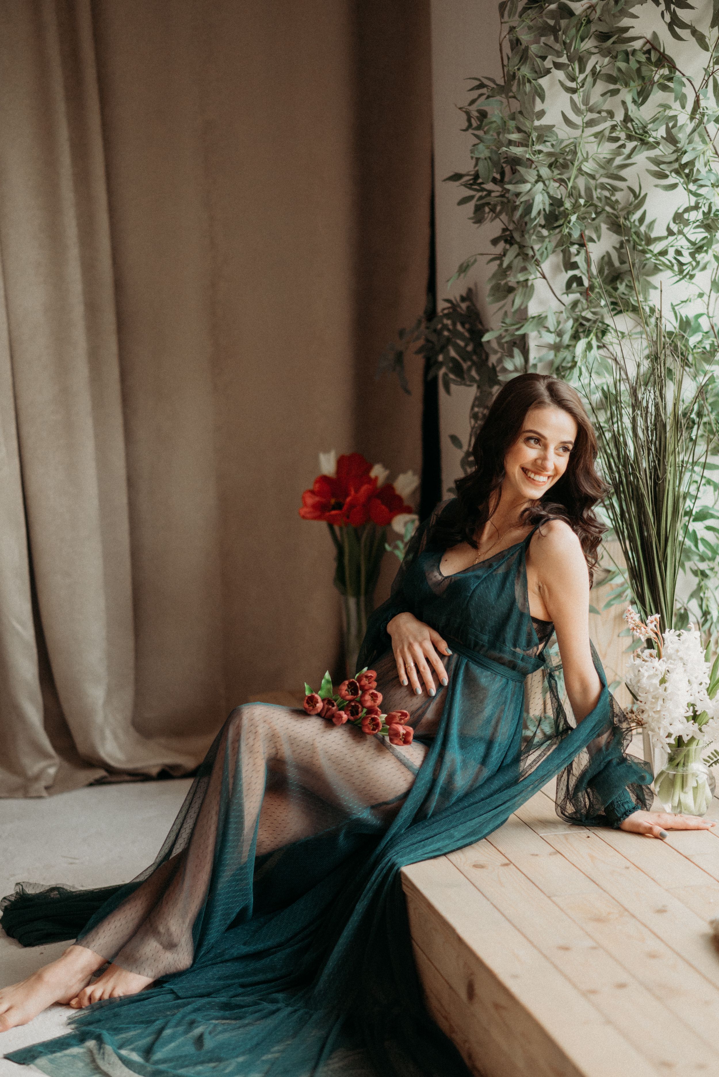 Elegant maternity session with flowing teal dress against floral backdrop.