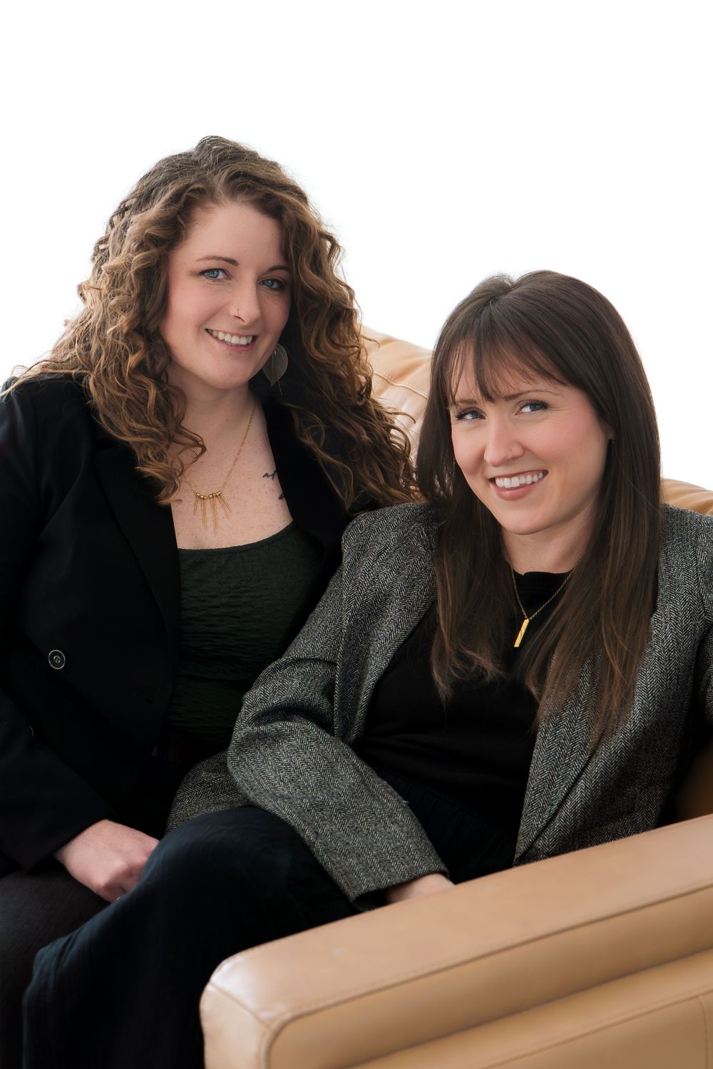 Two smiling women in blazers sitting together on a beige leather sofa against a white background.