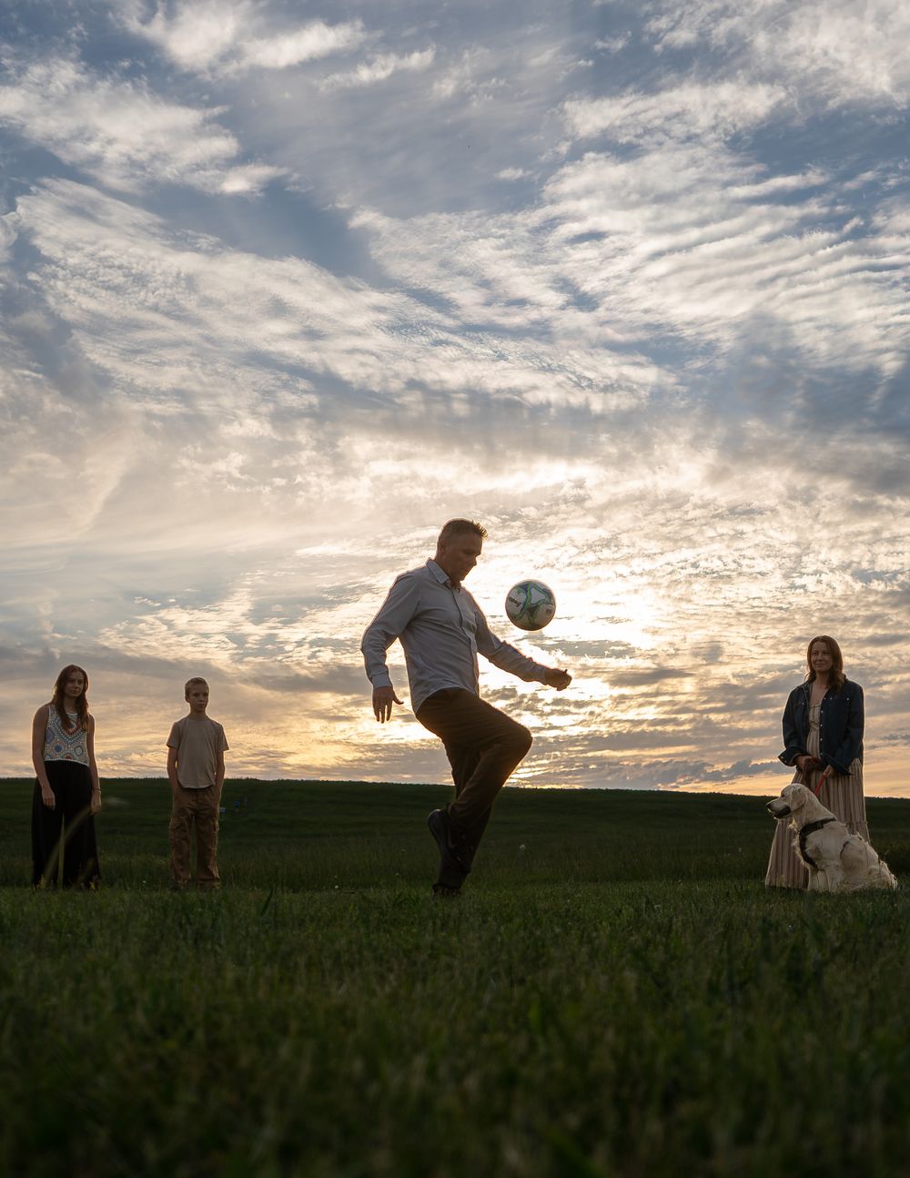 Family playing soccer on a green field at sunset with a dog nearby under a dramatic cloudy sky.
