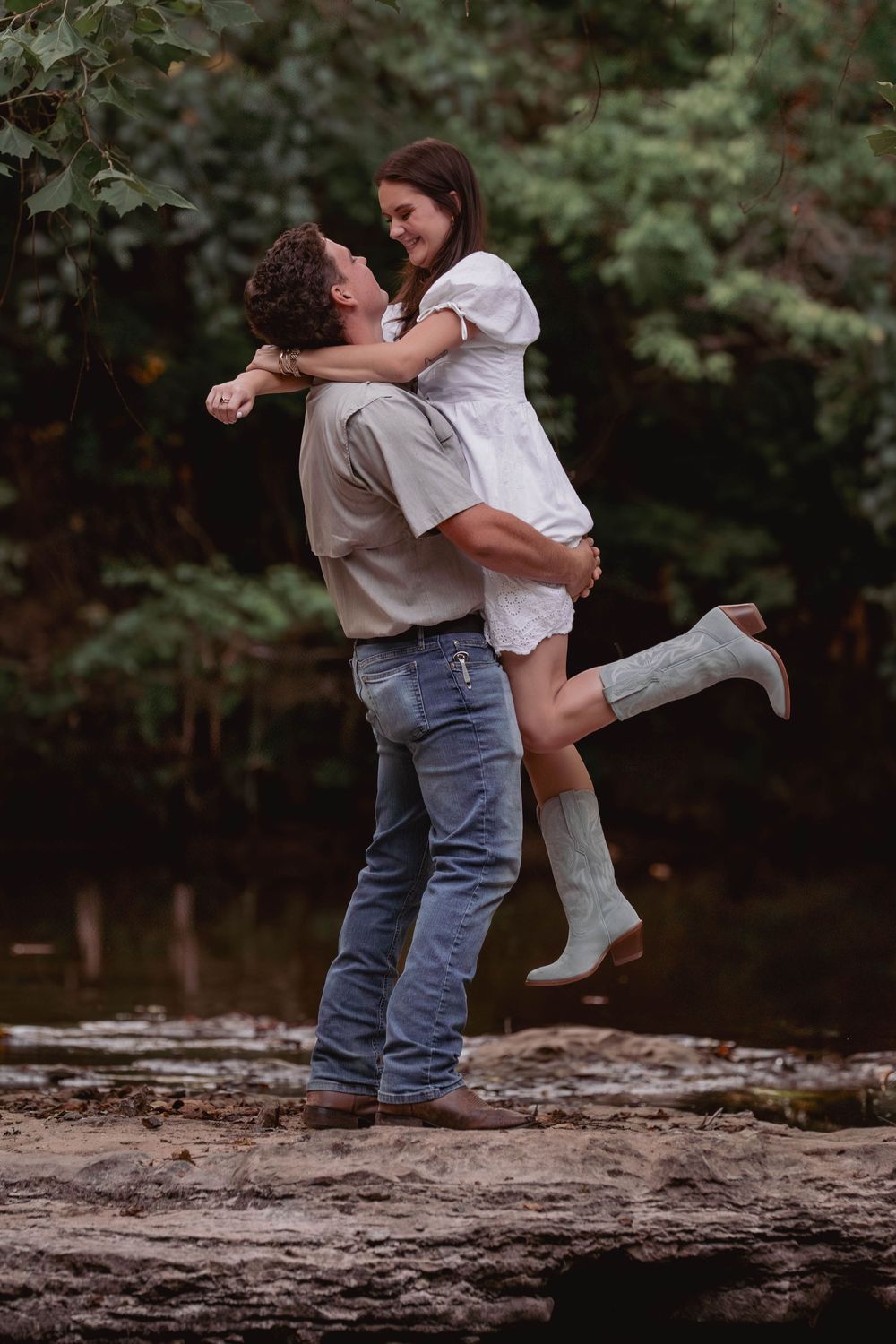 A romantic couple embraces on a rocky riverbank, with the woman wearing white boots lifted up by her partner in jeans.