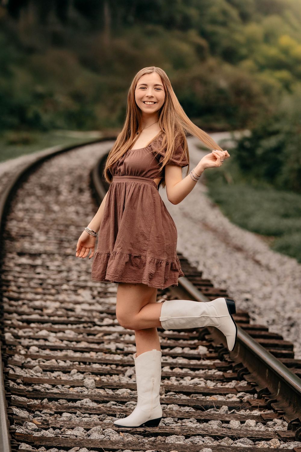 A person in a brown dress and white boots playfully dances along old railroad tracks in a wooded setting.