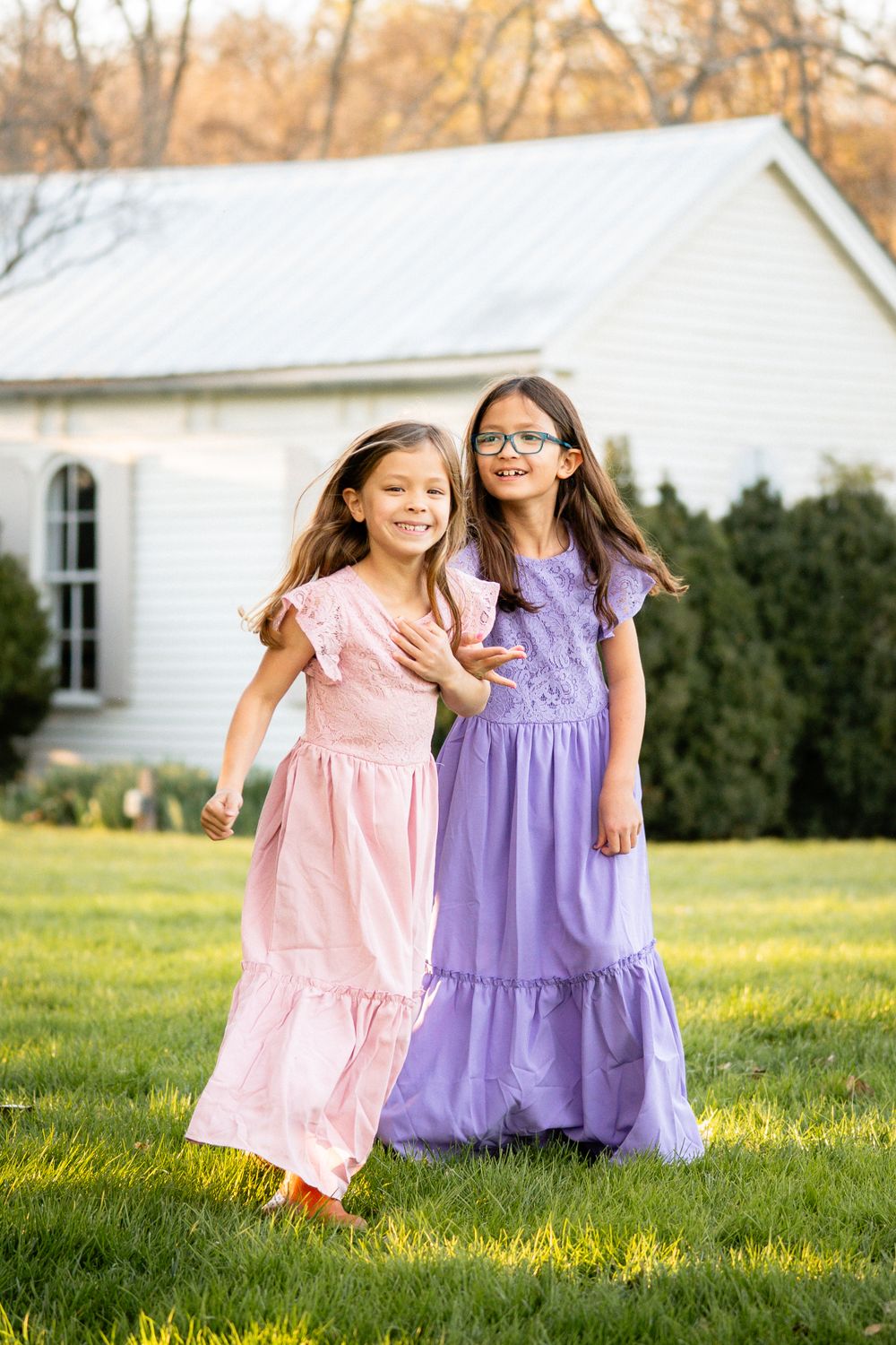 Two young children in pink and purple maxi dresses playing on a lawn in front of a white country church.