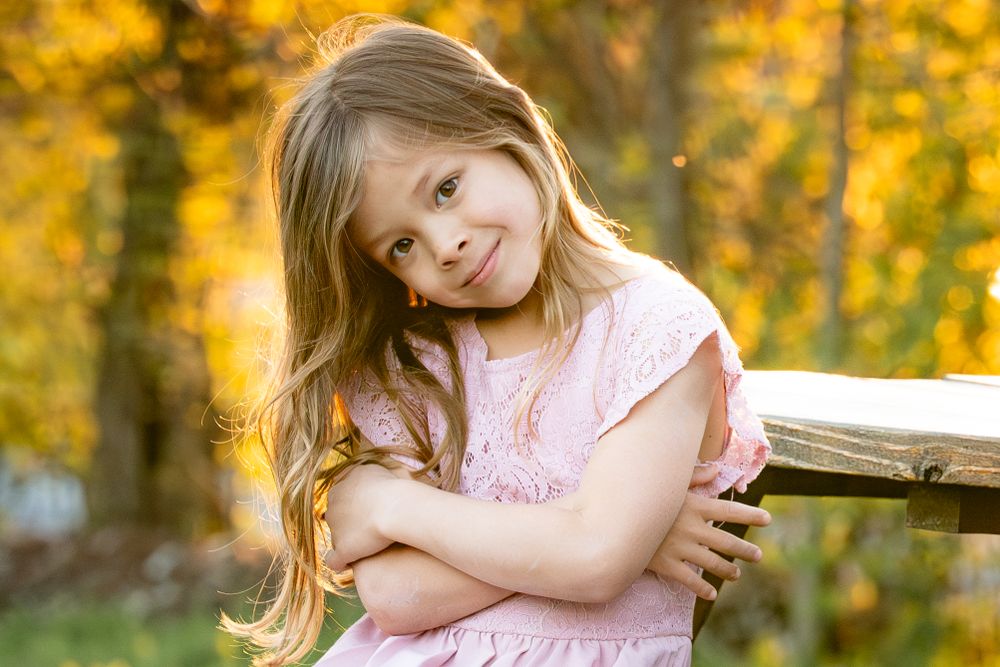 Young child in pink dress leaning against wooden bench in autumn setting with golden foliage background.