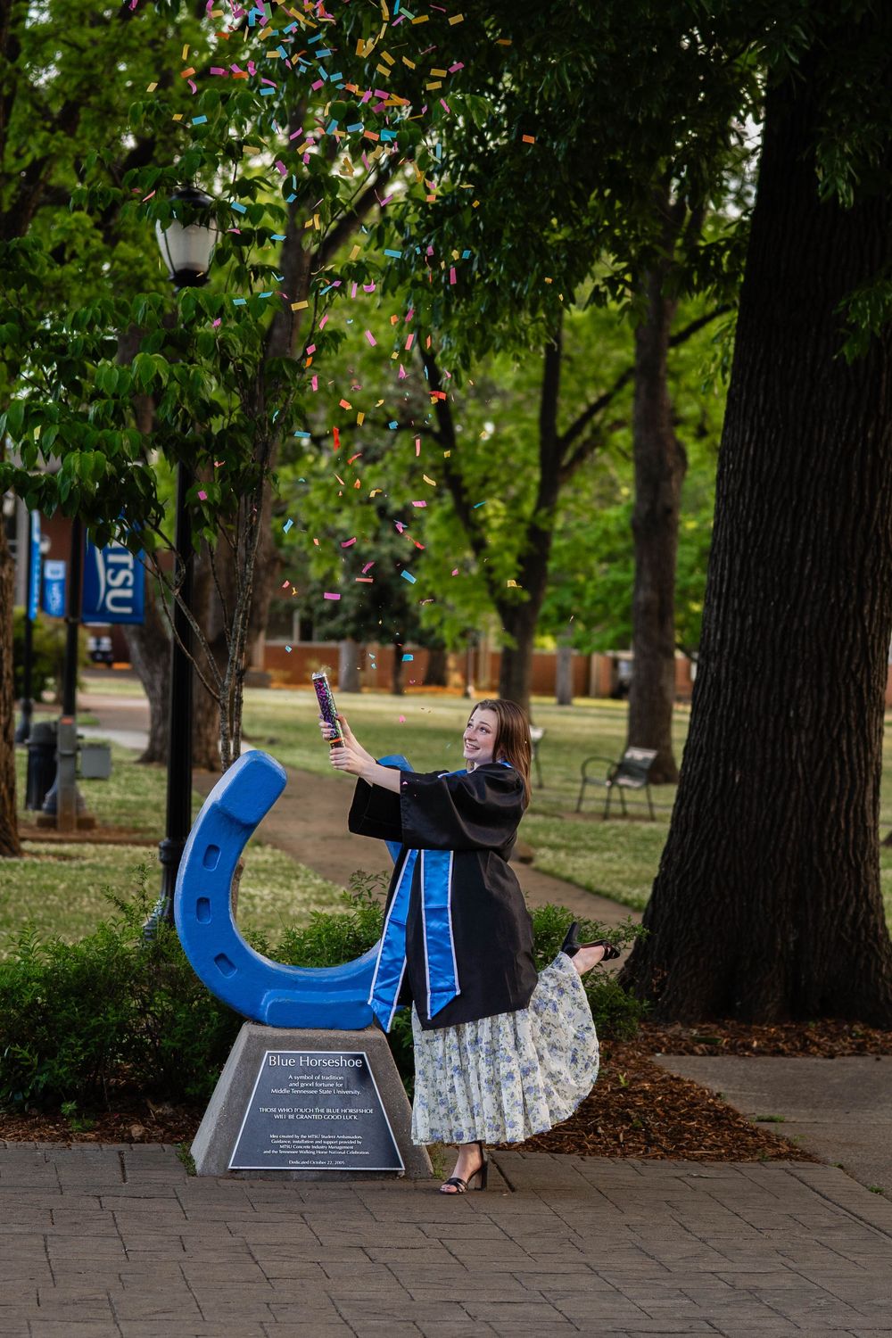 Graduate in cap and gown poses with blue horseshoe sculpture on college campus surrounded by trees.
