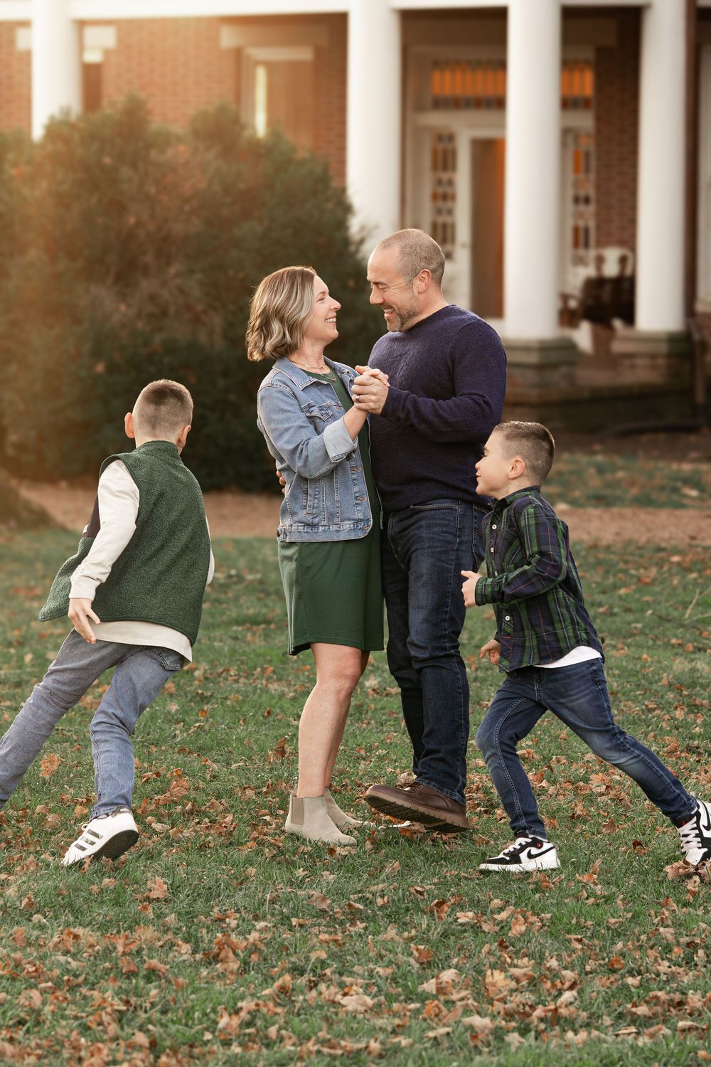 A playful sequence of family members chasing and running through autumn leaves on a lawn outside a building.