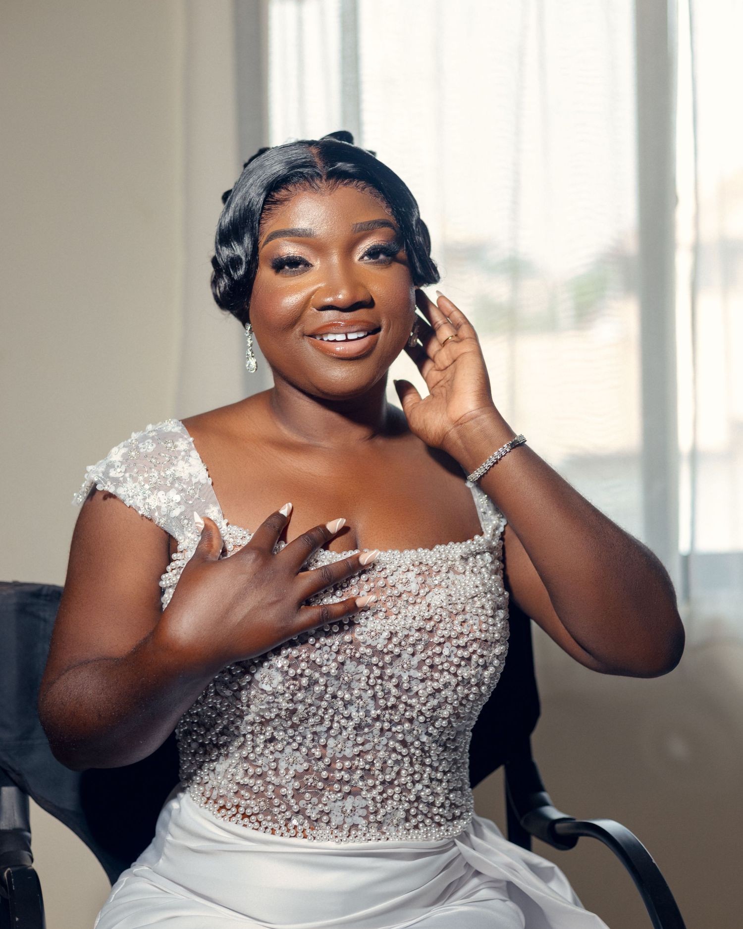 Bride in beaded white gown shows joyful expression while touching face and chest.
