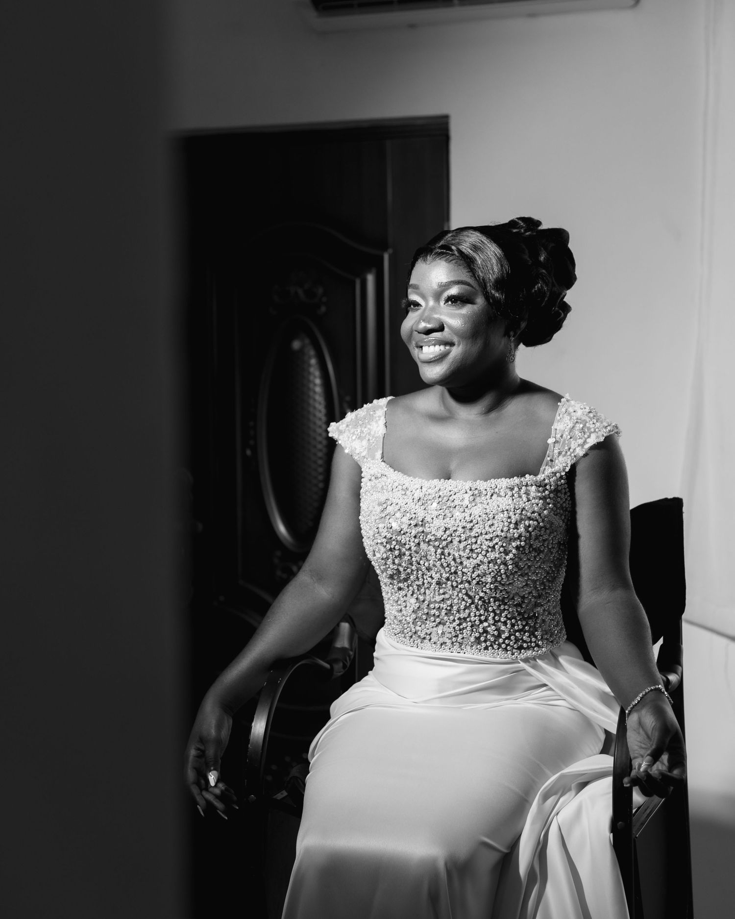 Bride sitting in elegant beaded white wedding dress with cap sleeves smiles radiantly in black and white photo.