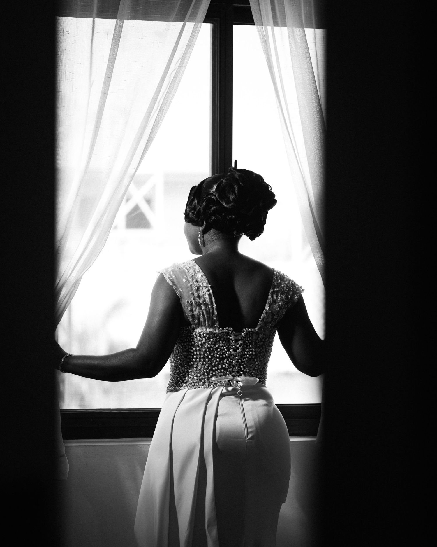Silhouetted bride in beaded dress gazes through window with flowing curtains.
