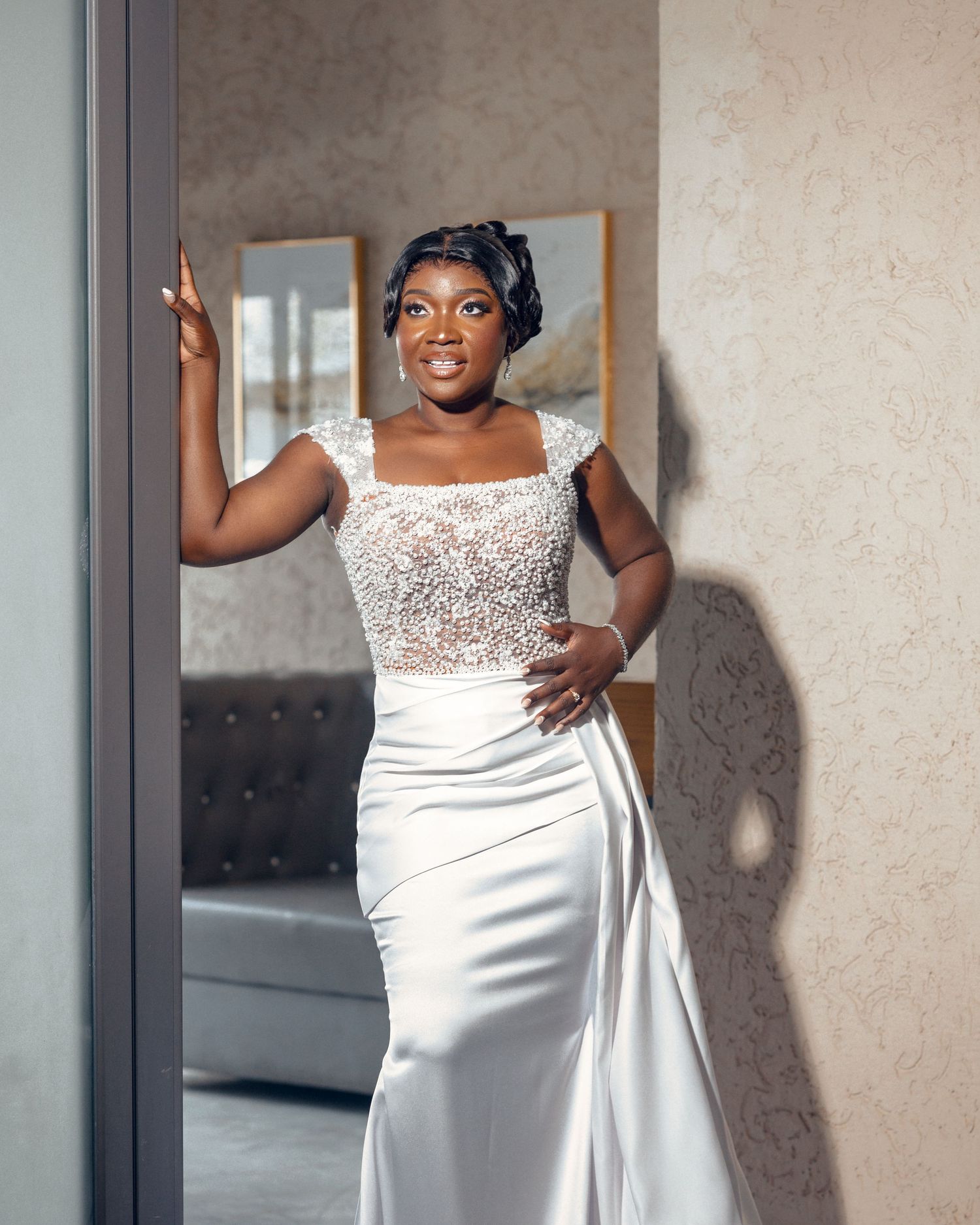 Bride in fitted white beaded gown poses elegantly against neutral wall.