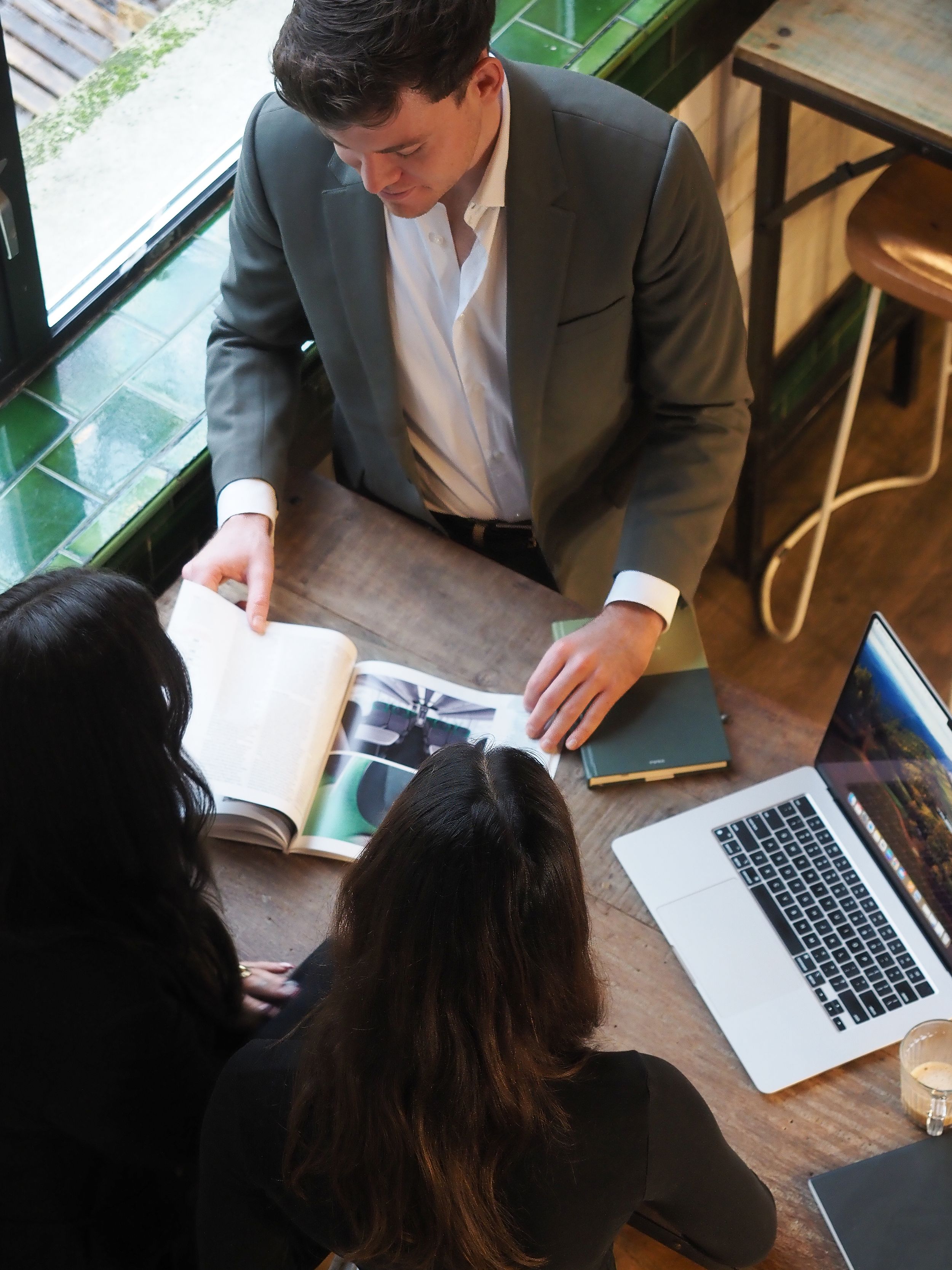 A businessman in a gray suit reviews an open book with two women at a wooden table with a laptop.