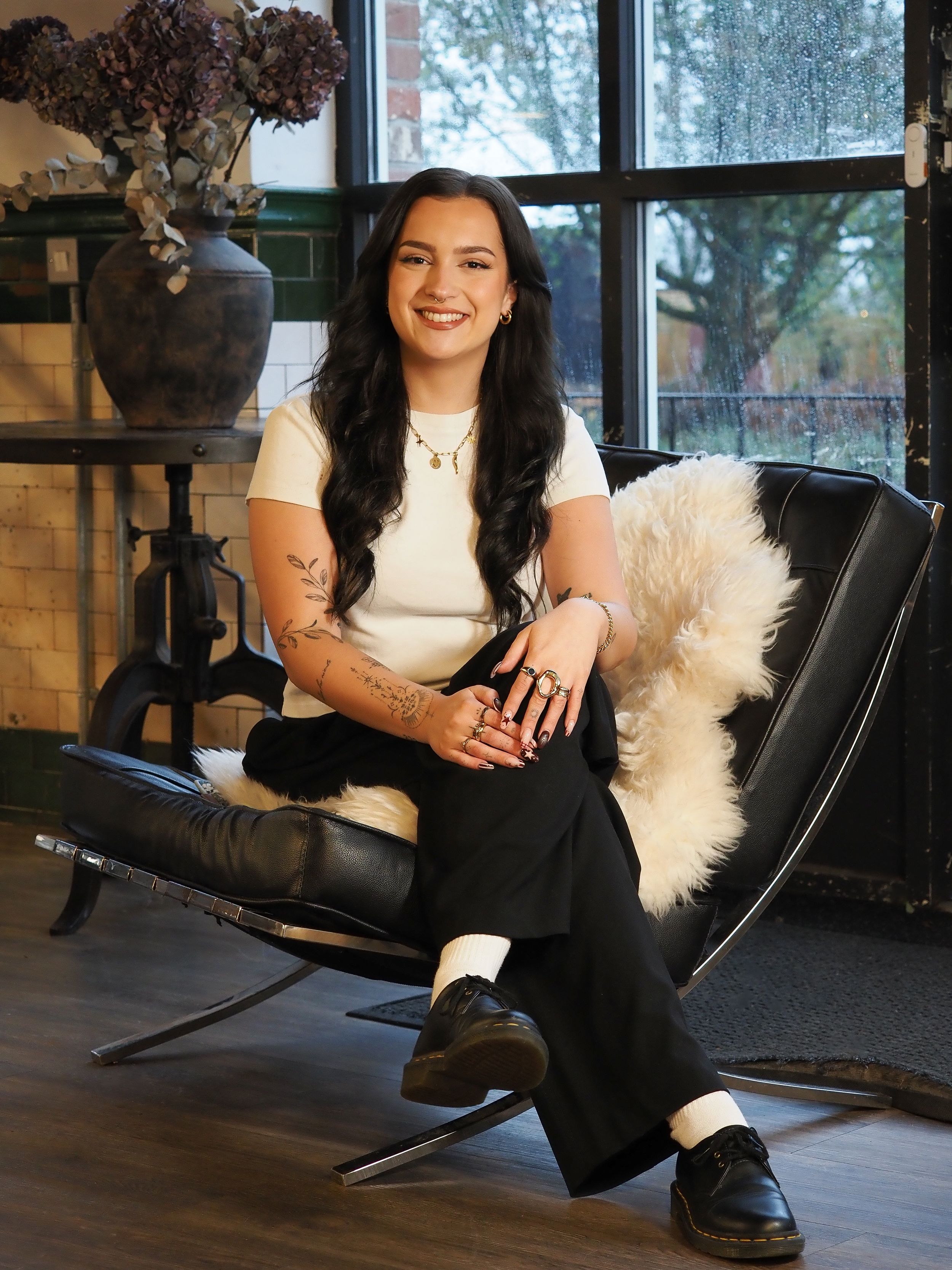 Young woman with long dark hair and tattoos sits smiling in a black leather chair wearing a white top and black pants.