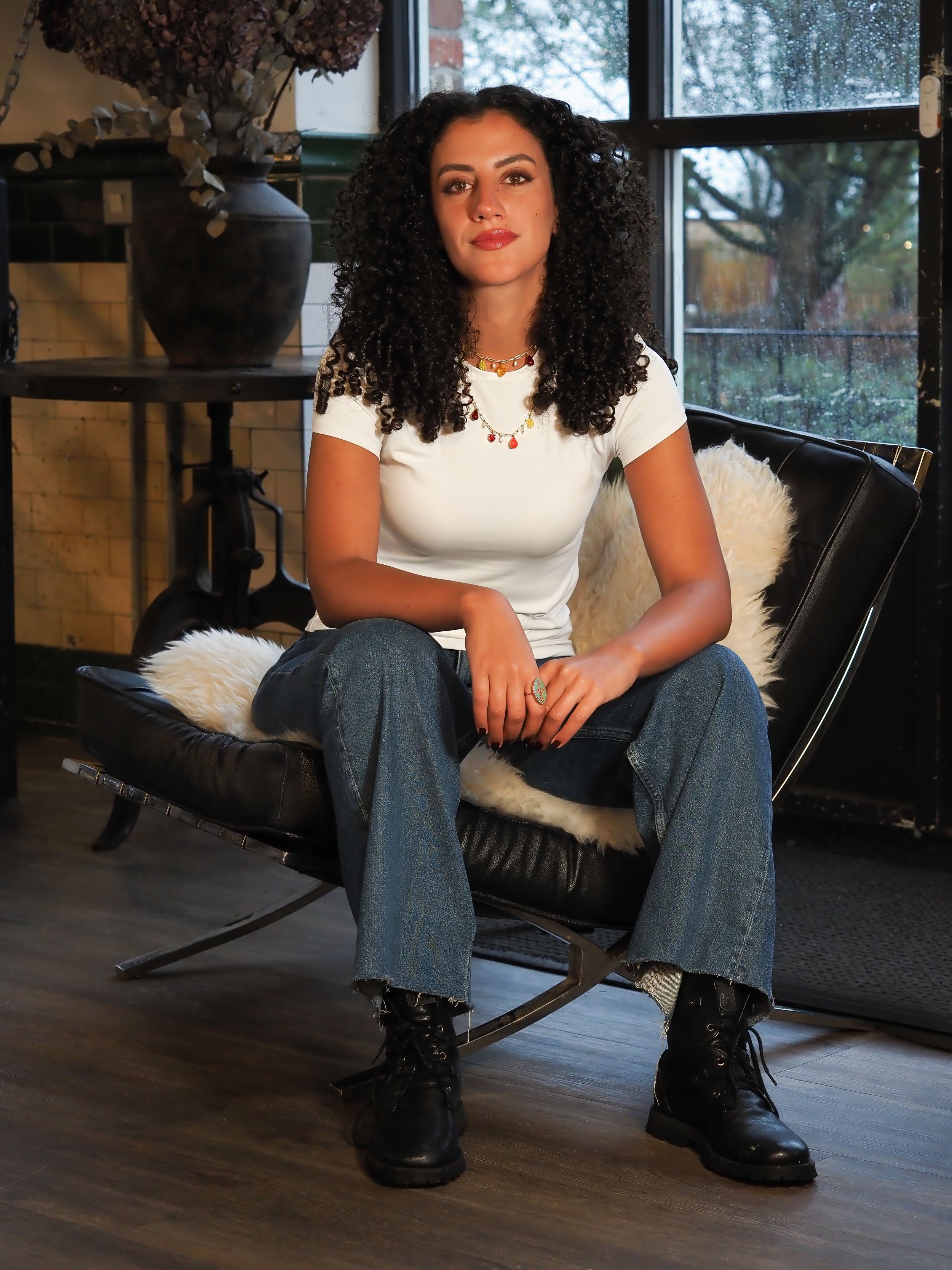 Young woman with curly hair sitting confidently in a black leather chair wearing jeans and boots.