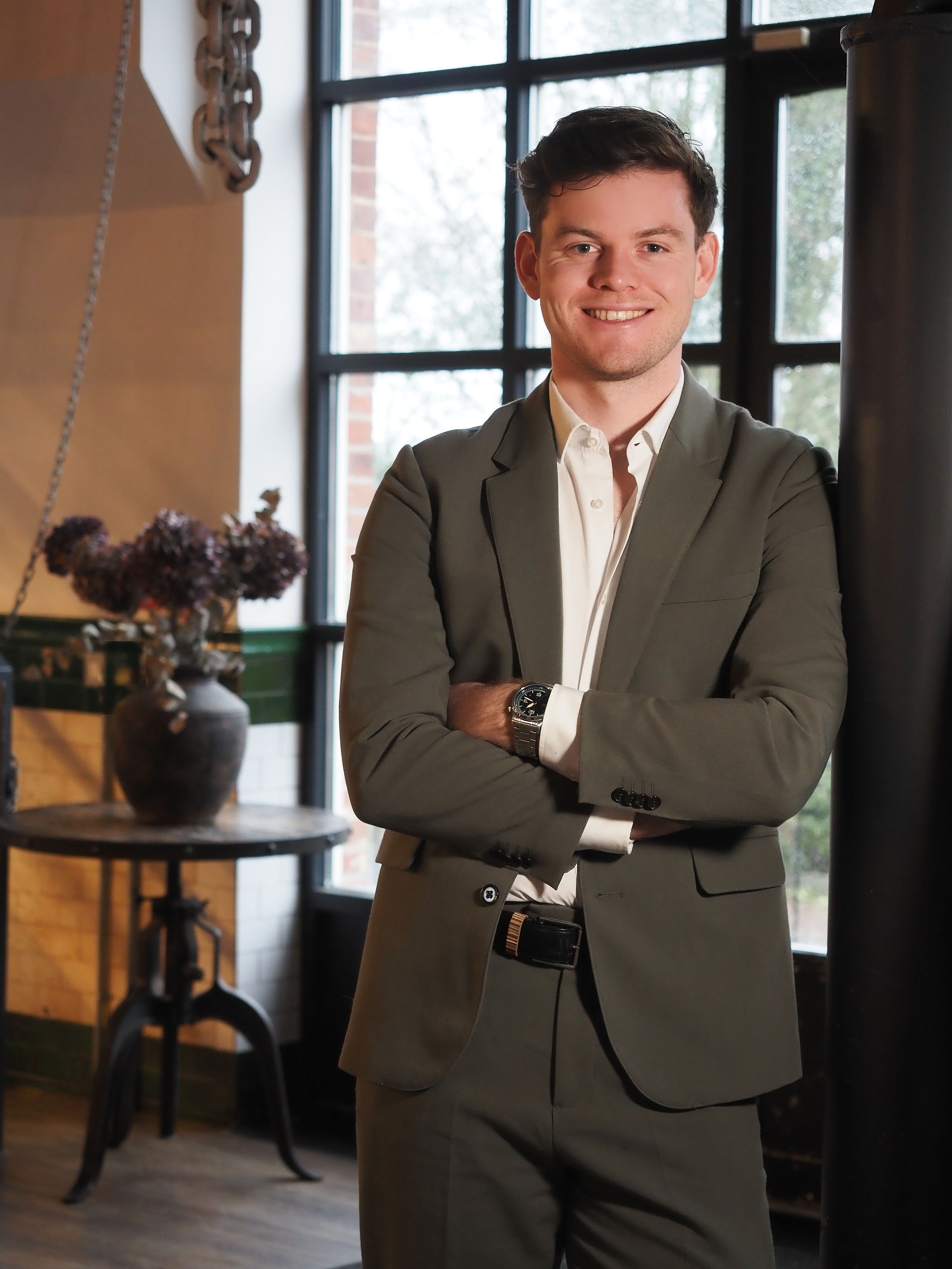 Young professional man in olive green suit smiling with arms crossed in modern interior setting.