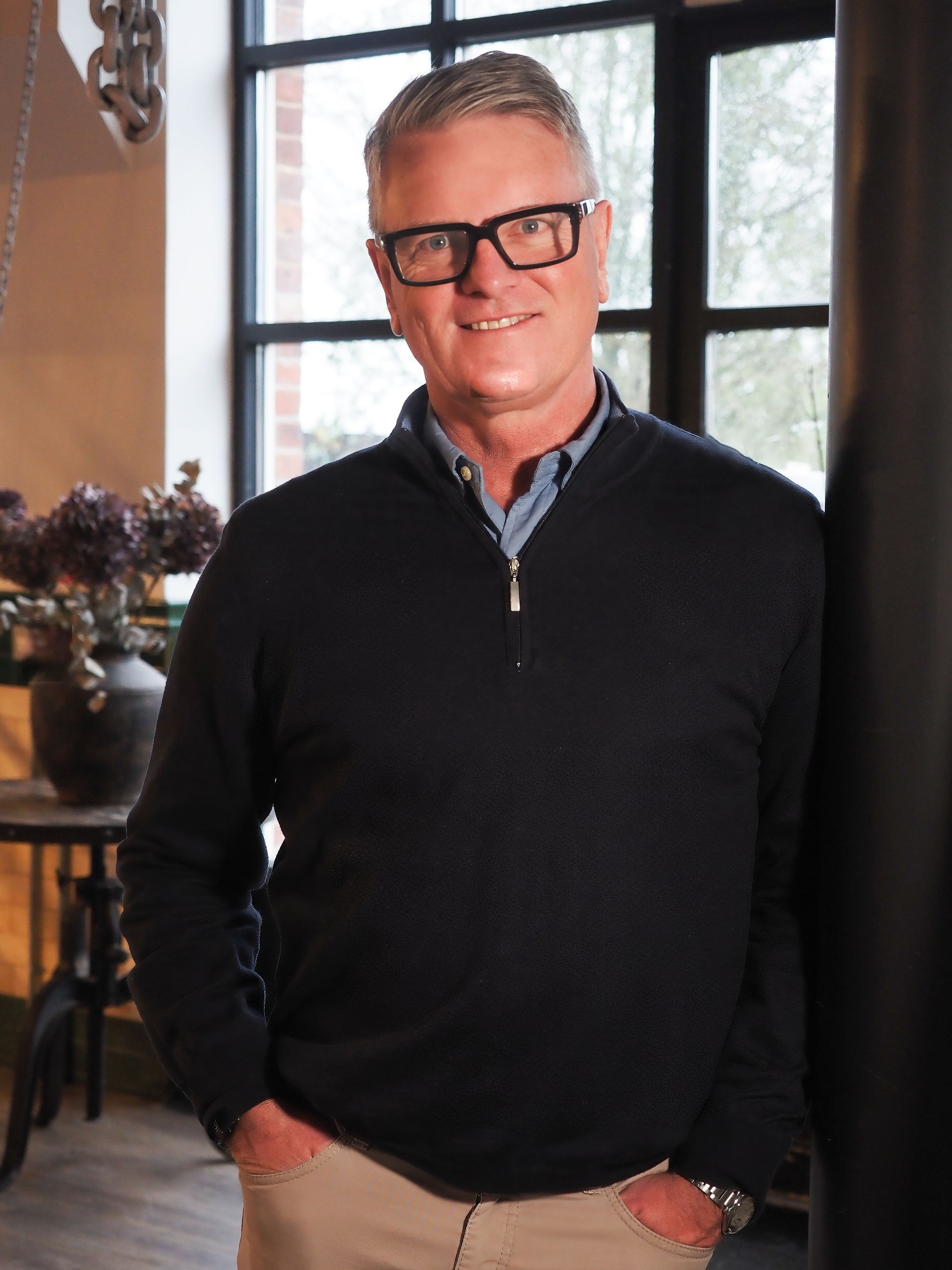 Smiling middle-aged man with glasses wearing a black quarter-zip sweater standing in a modern interior space.