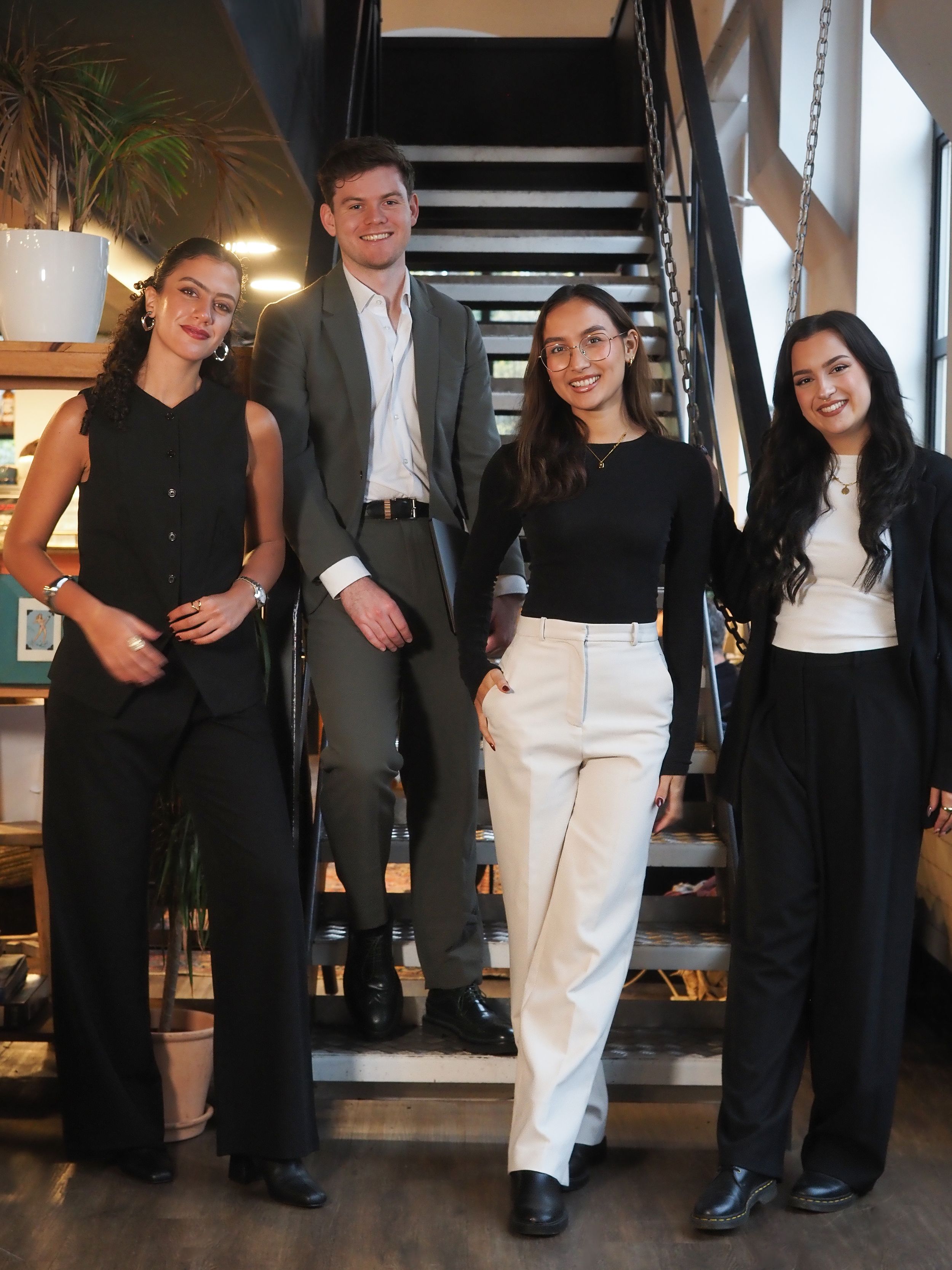 Four young professionals posing together on stairs in a modern interior, dressed in smart casual black and neutral outfits.