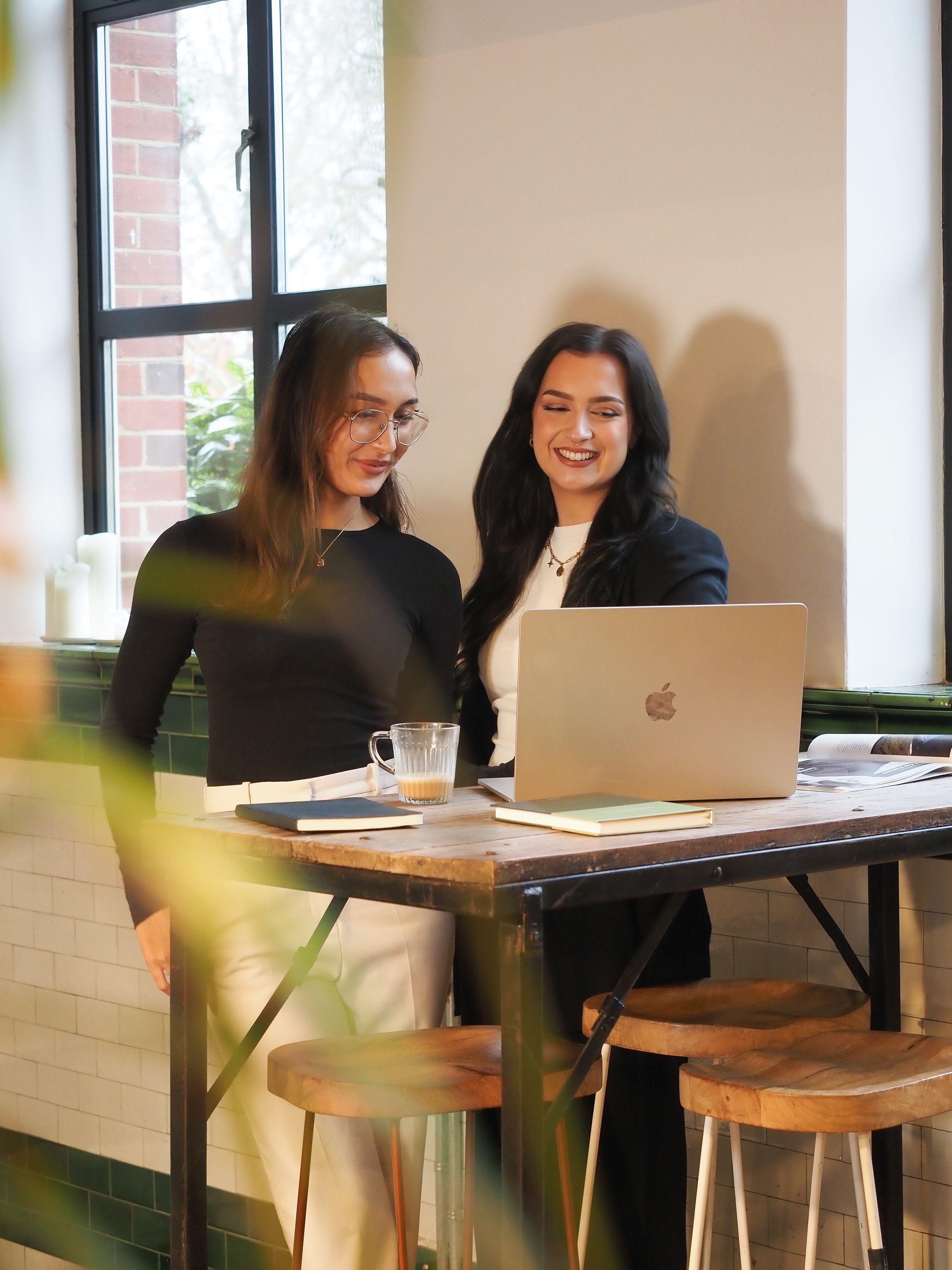 Two professional women collaborating on a MacBook laptop at a wooden standing desk in a modern office.