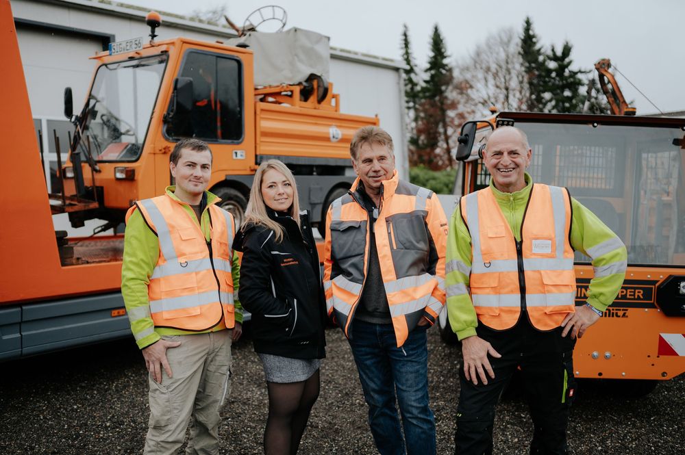Arbeiter in gut sichtbarer Sicherheitskleidung stehen neben orangefarbenen Wartungsfahrzeugen auf einer Baustelle.
