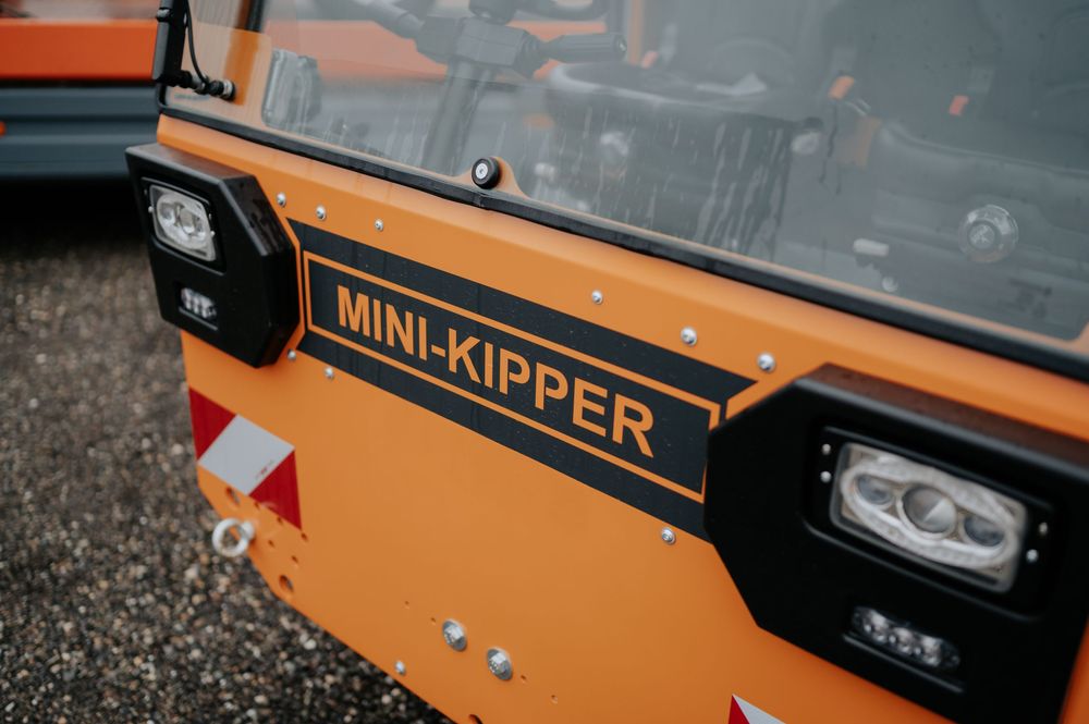 Orangefarbener Mini-Kipper mit schwarzer Zierleiste und reflektierenden Sicherheitsmarkierungen, geparkt auf Kiesfläche.