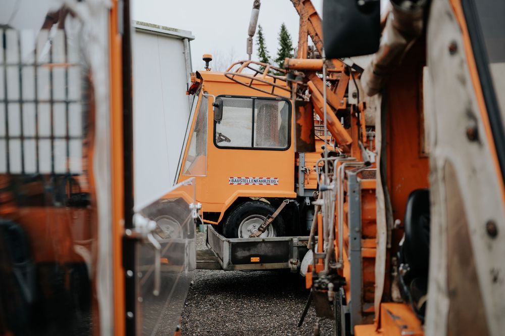 Orangefarbene Baumaschinen und -geräte stehen auf einer Baustelle mit Metallzäunen im Hintergrund in einer Reihe.
