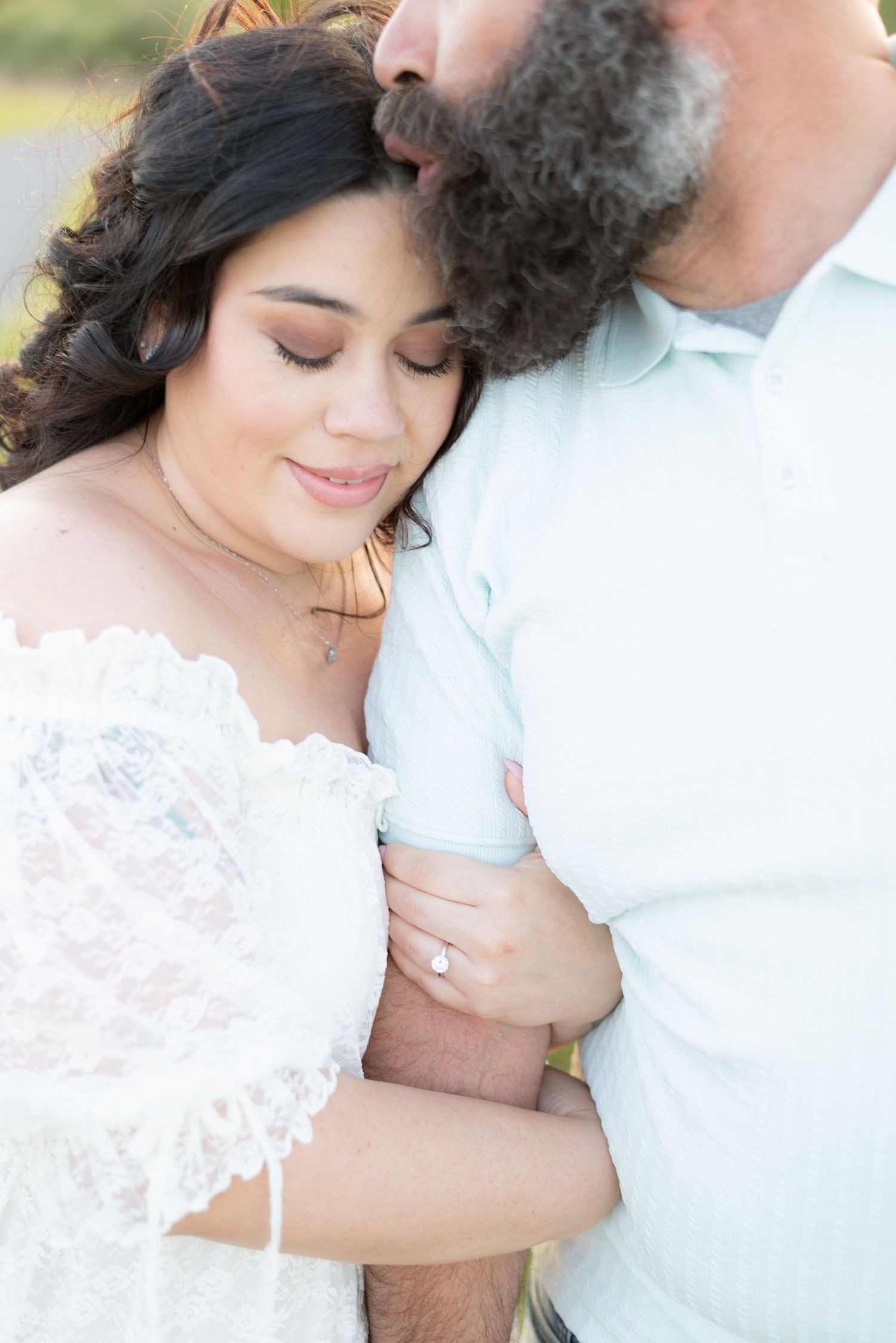 Romantic couple embracing outdoors, woman in white lace dress with engagement ring, man kissing her head tenderly.