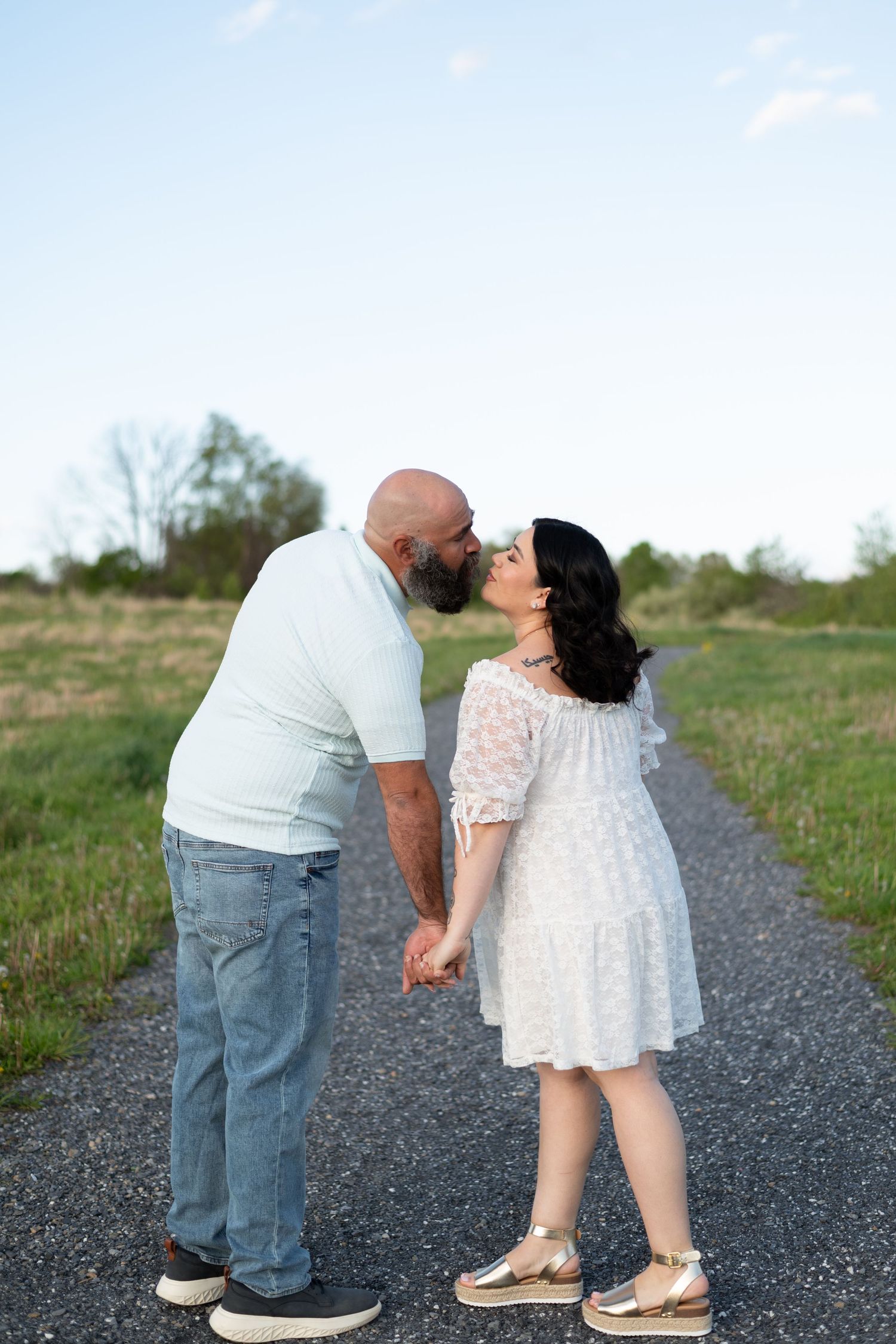 A couple shares a romantic kiss while holding hands on a scenic path, man in white shirt and jeans, woman in white lace dress.
