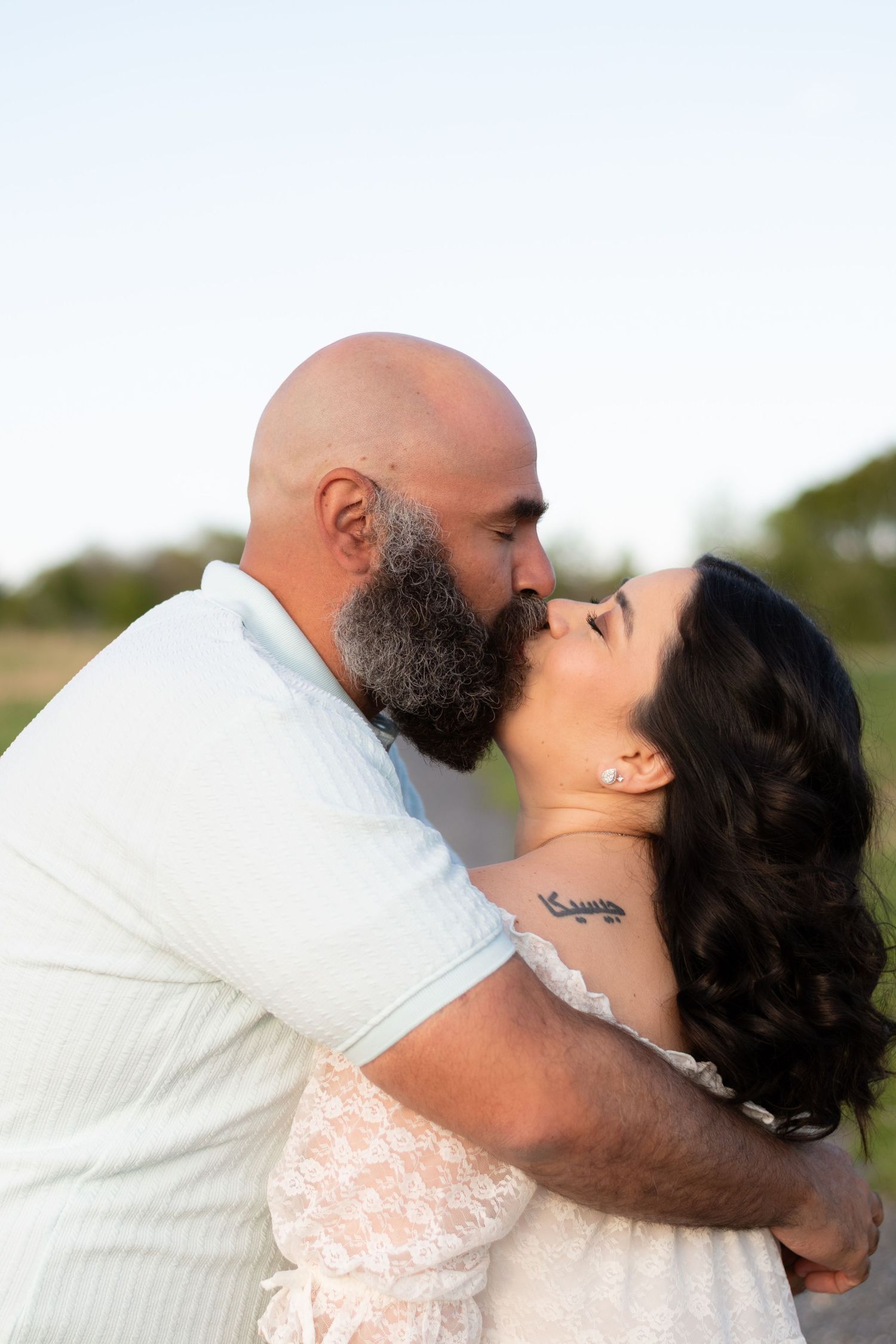 Bald bearded man kissing dark-haired woman with tattoo outdoors during golden hour engagement photo.