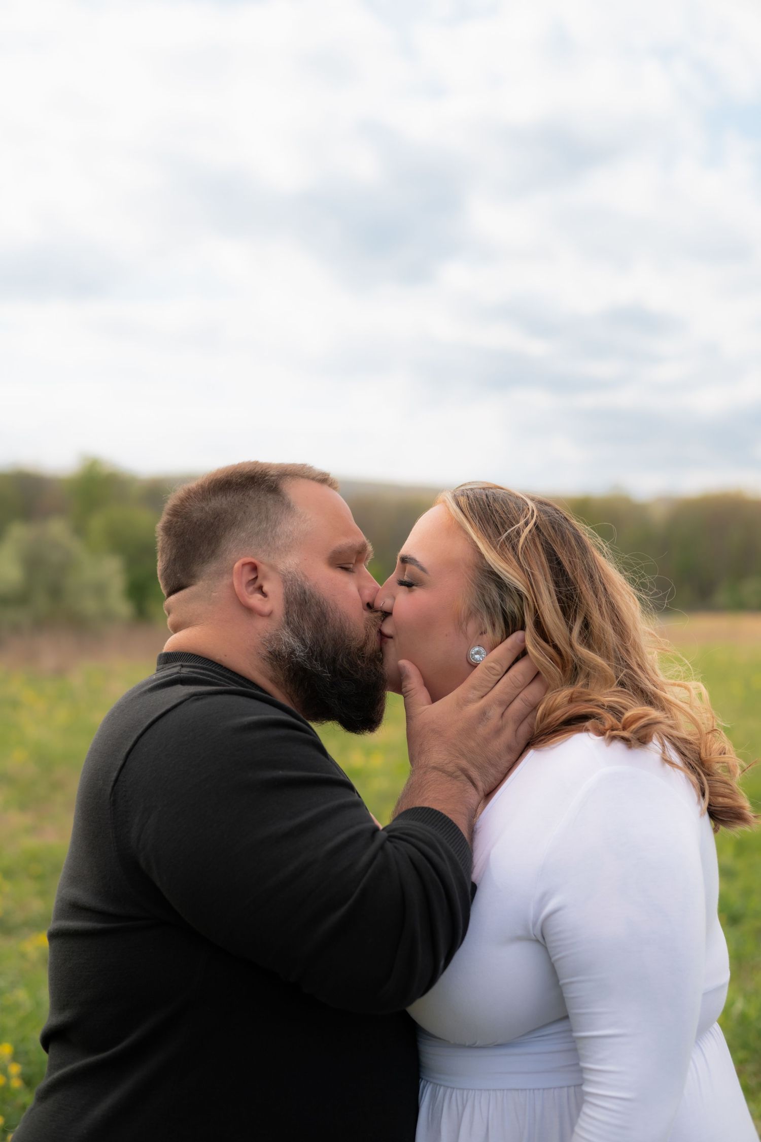 A couple shares a romantic kiss in a green field, the man in black and woman in white, under a cloudy sky.