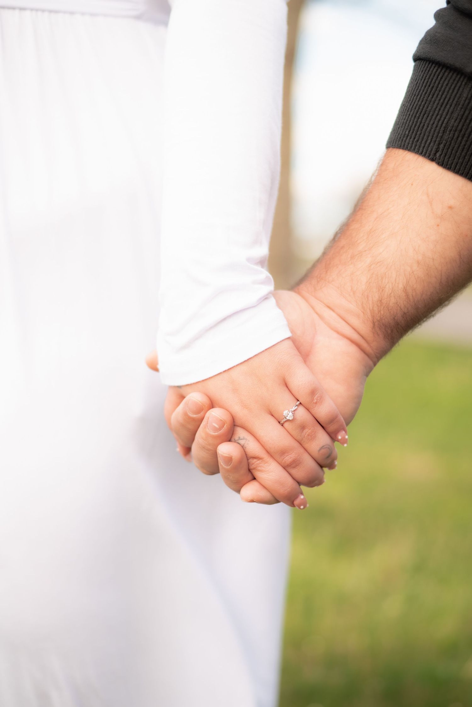 Close-up of a couple holding hands, woman wearing an engagement ring, dressed in white, with green background.