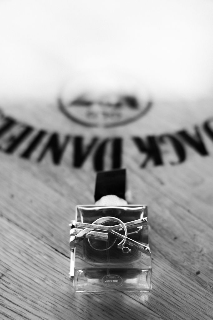 Black and white photo of YSL cologne bottle on wood surface with Jack Daniels barrel in background.