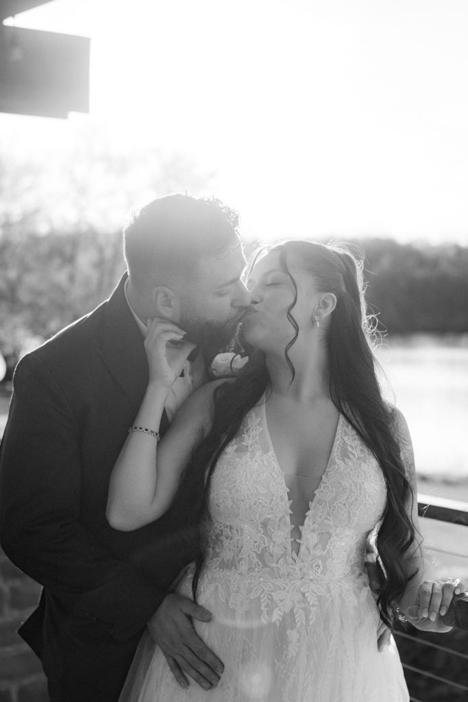 Black and white photo of a bride and groom sharing a romantic kiss, backlit by golden sunlight outdoors.