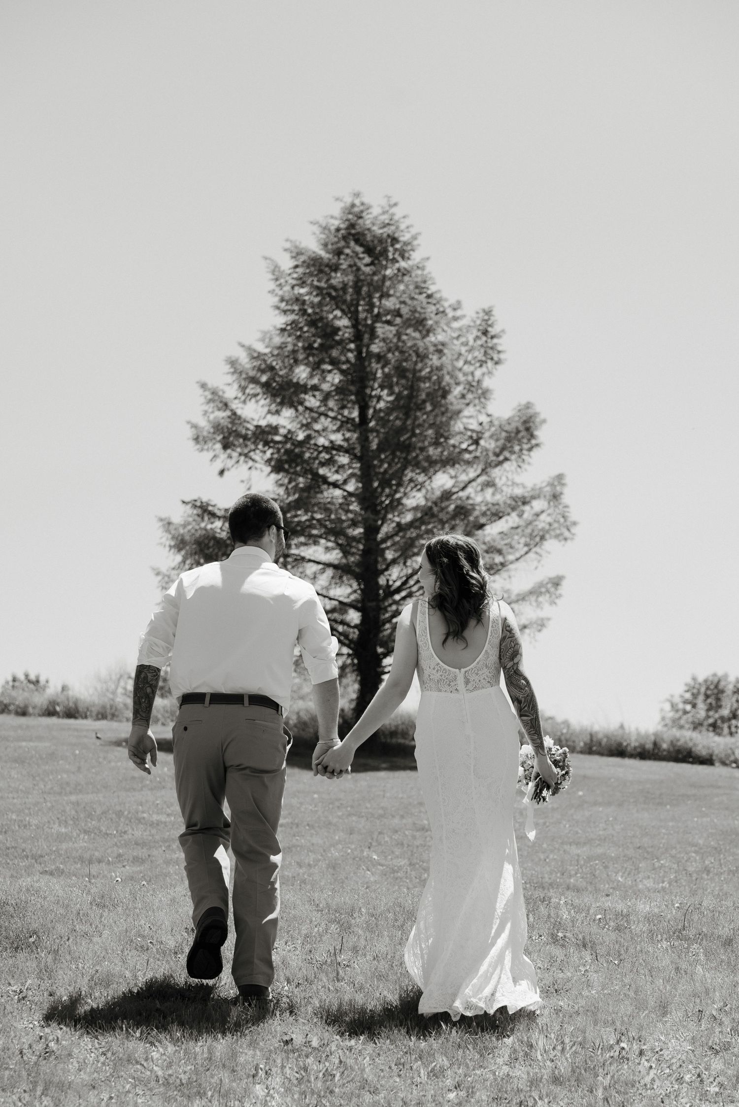 Black and white photo of a bride and groom holding hands walking through a sunny field toward a tall tree.