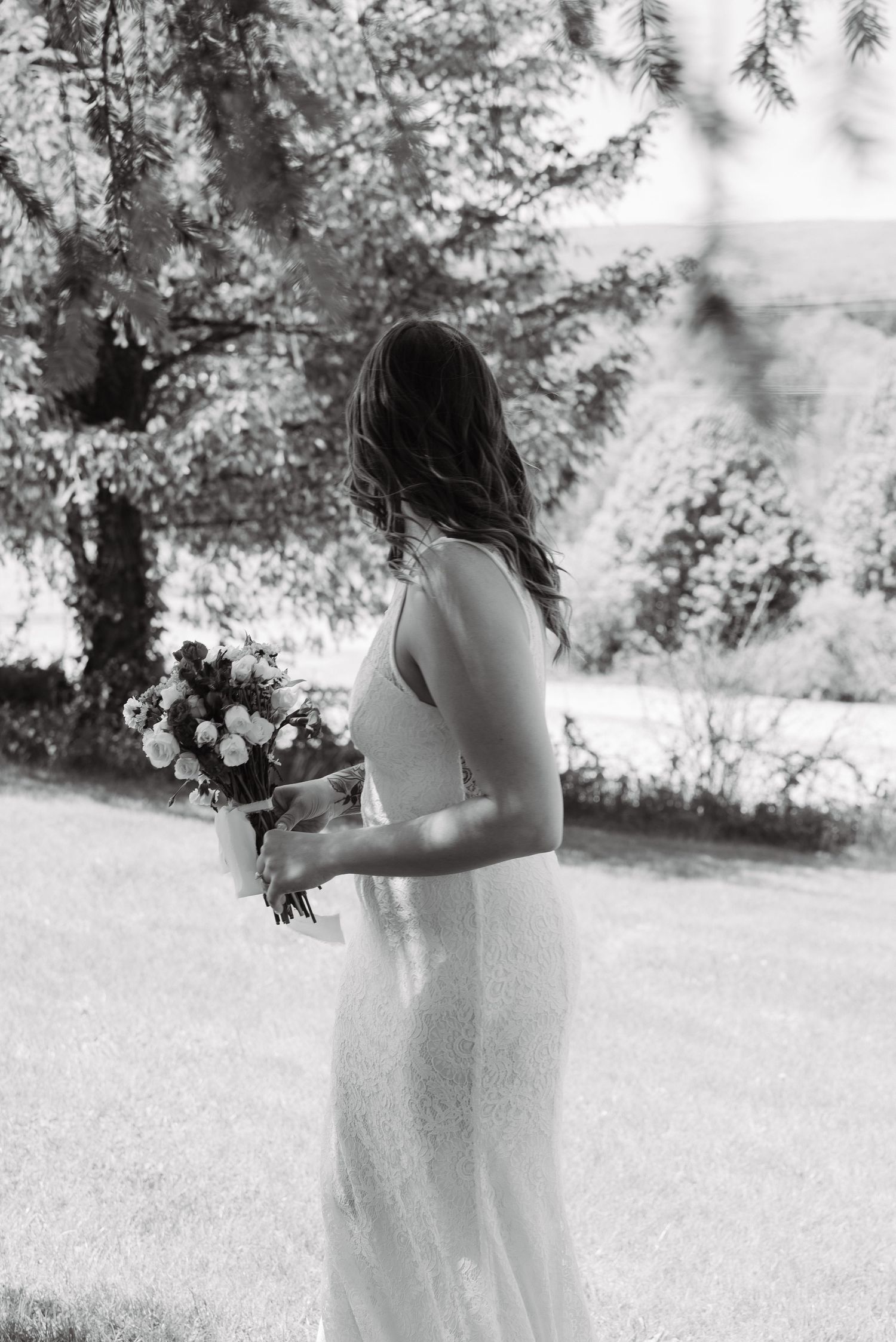 Black and white photo of a bride in a sleek gown holding a bouquet, standing outdoors with trees in the background.