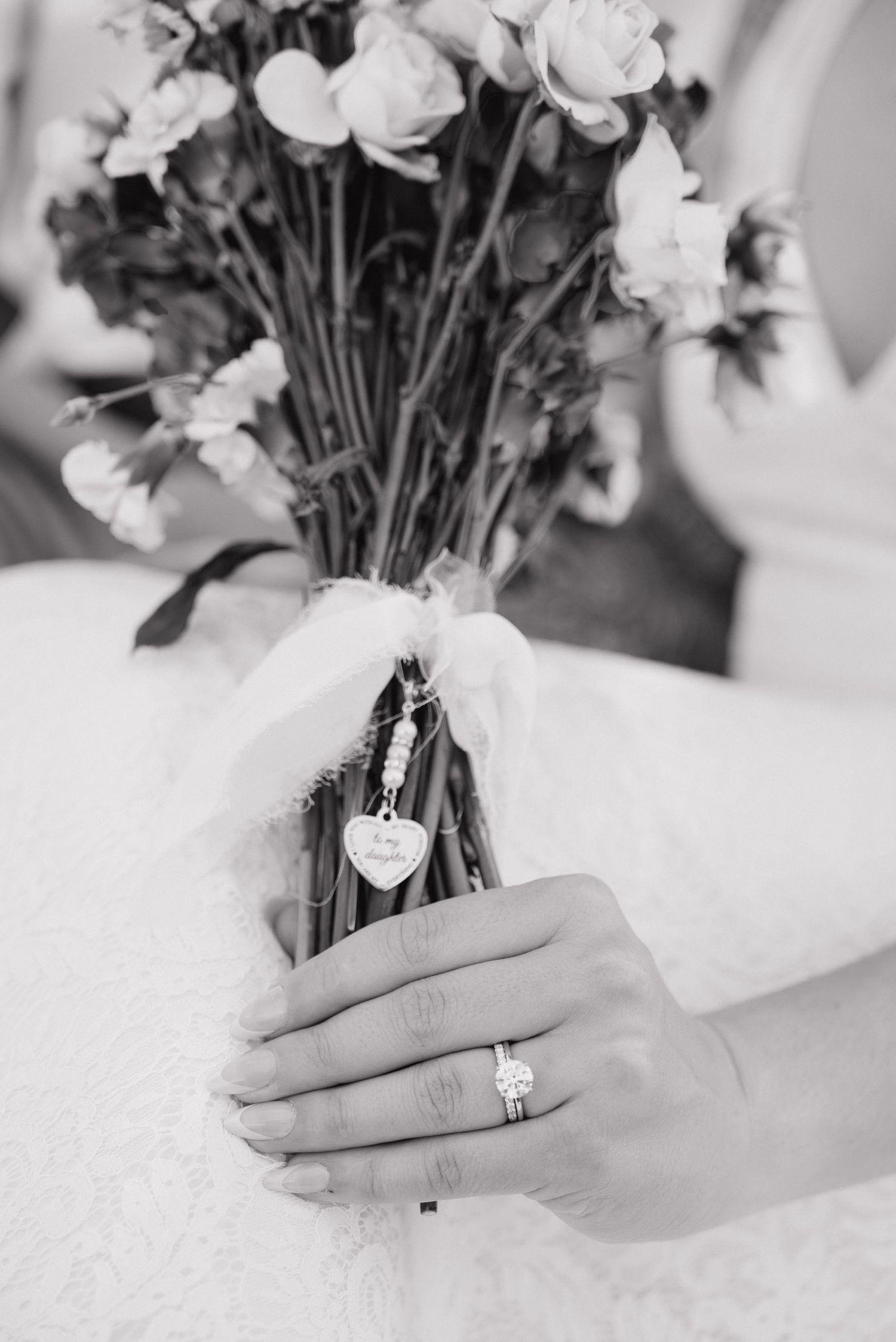Black and white photo of bride holding rose bouquet with heart charm, showcasing elegant diamond engagement ring.