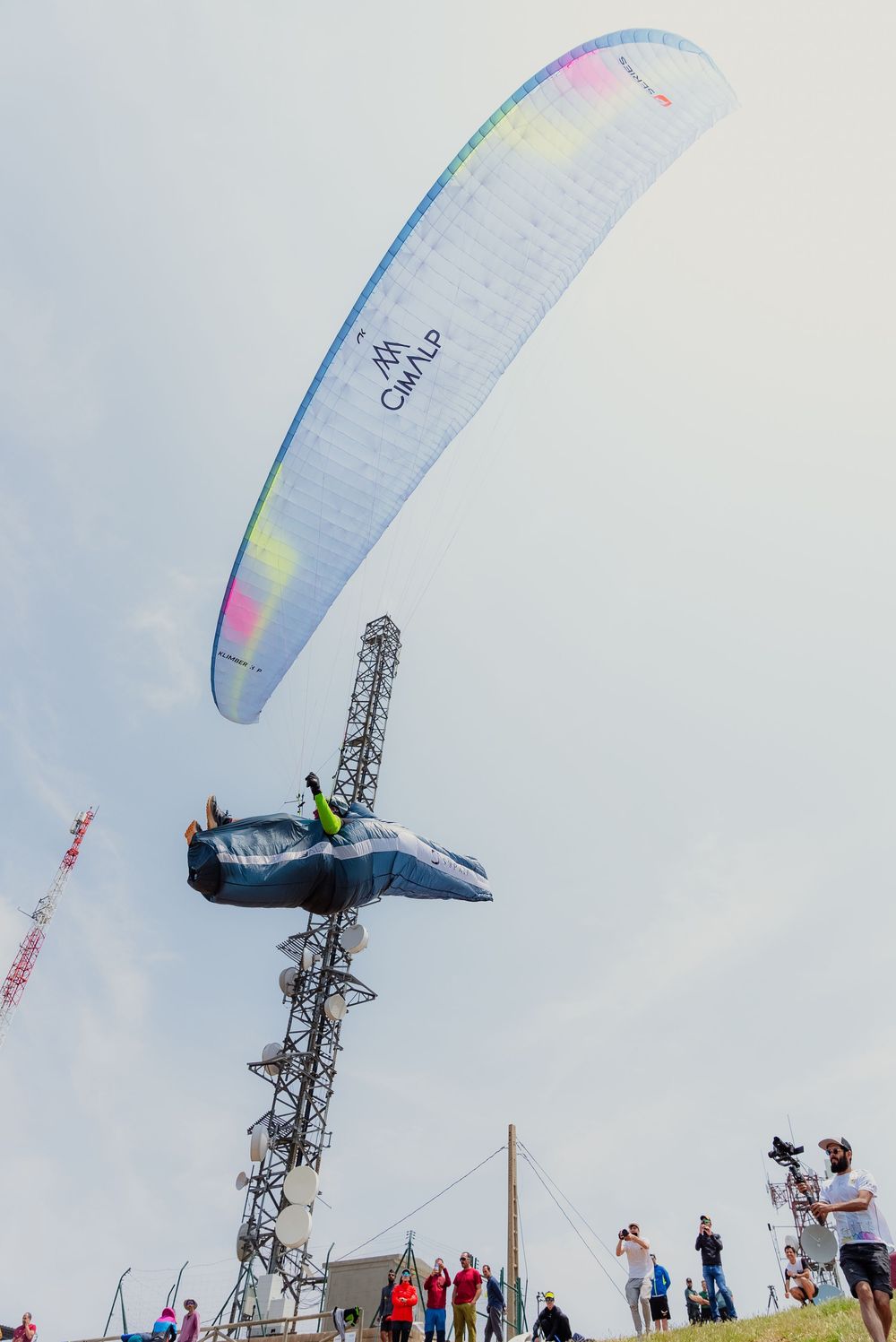 A paraglider soars past a tall communications tower as spectators watch from below against a cloudy sky.