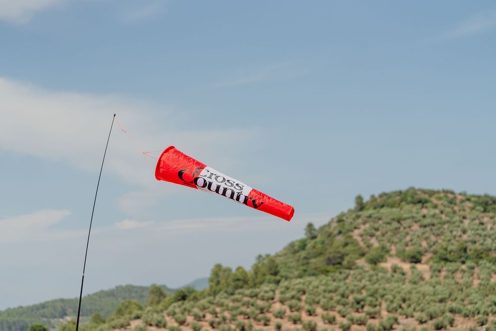 Red wind sock blows in the breeze against blue sky and green hillside landscape.