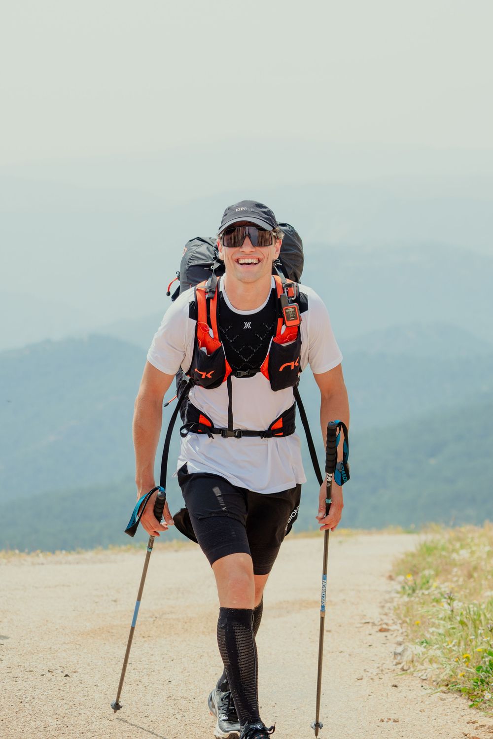 Hiker with trekking poles and backpack climbs uphill on a dusty mountain trail during a sunny day.